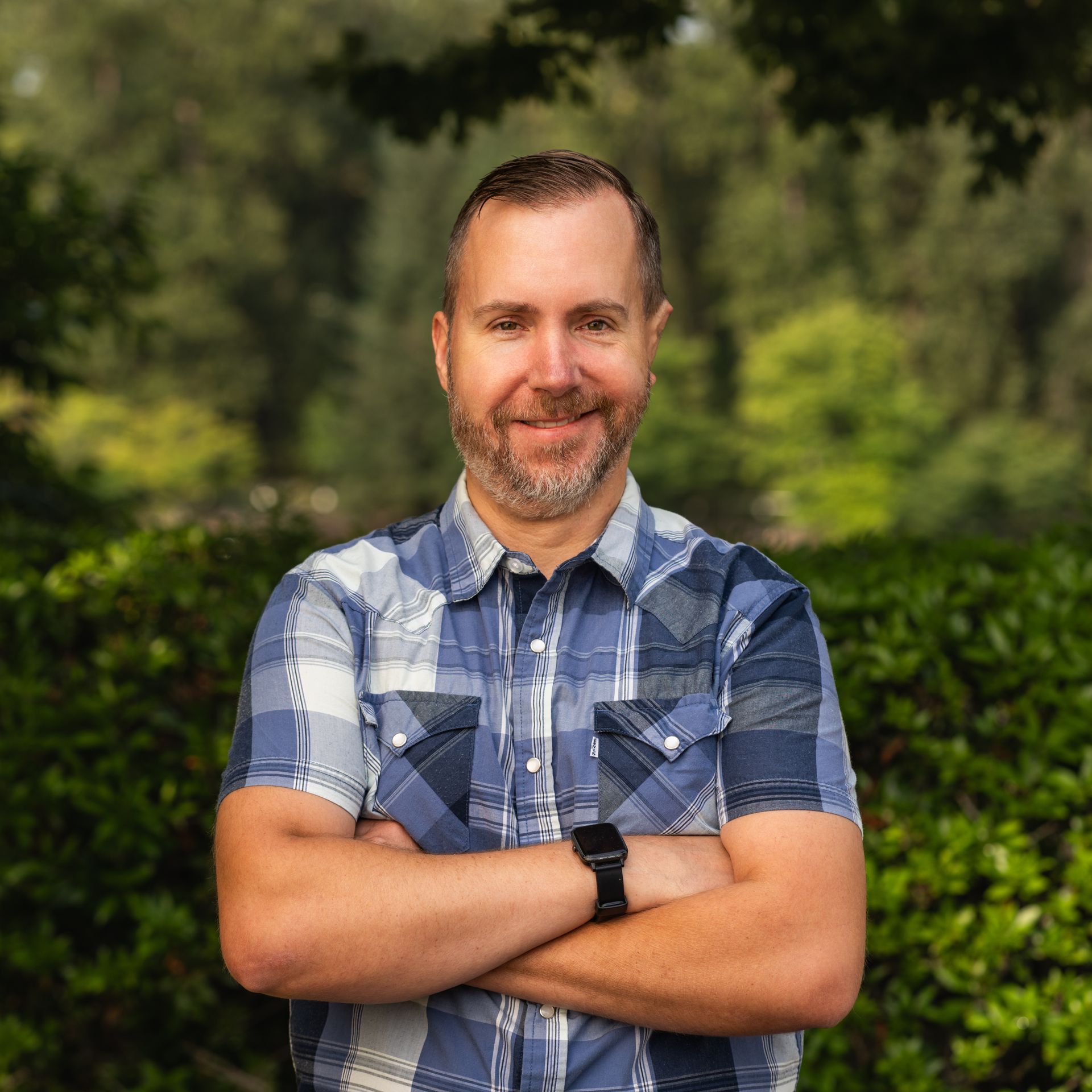 Man with arms crossed, wearing blue plaid shirt and watch, smiling in front of green foliage.