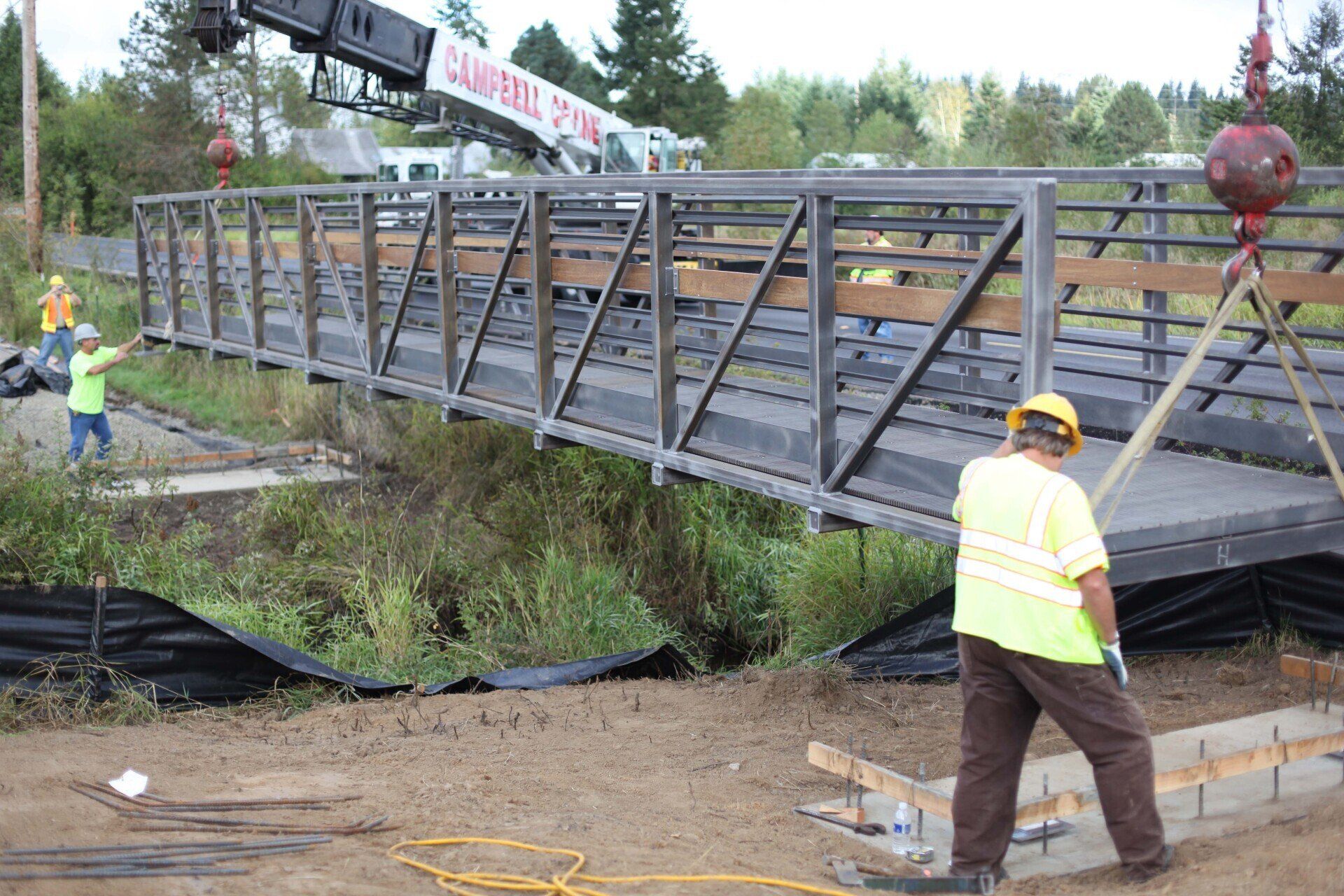 Workers install a metal bridge over a ditch, using a crane. One worker guides the bridge.
