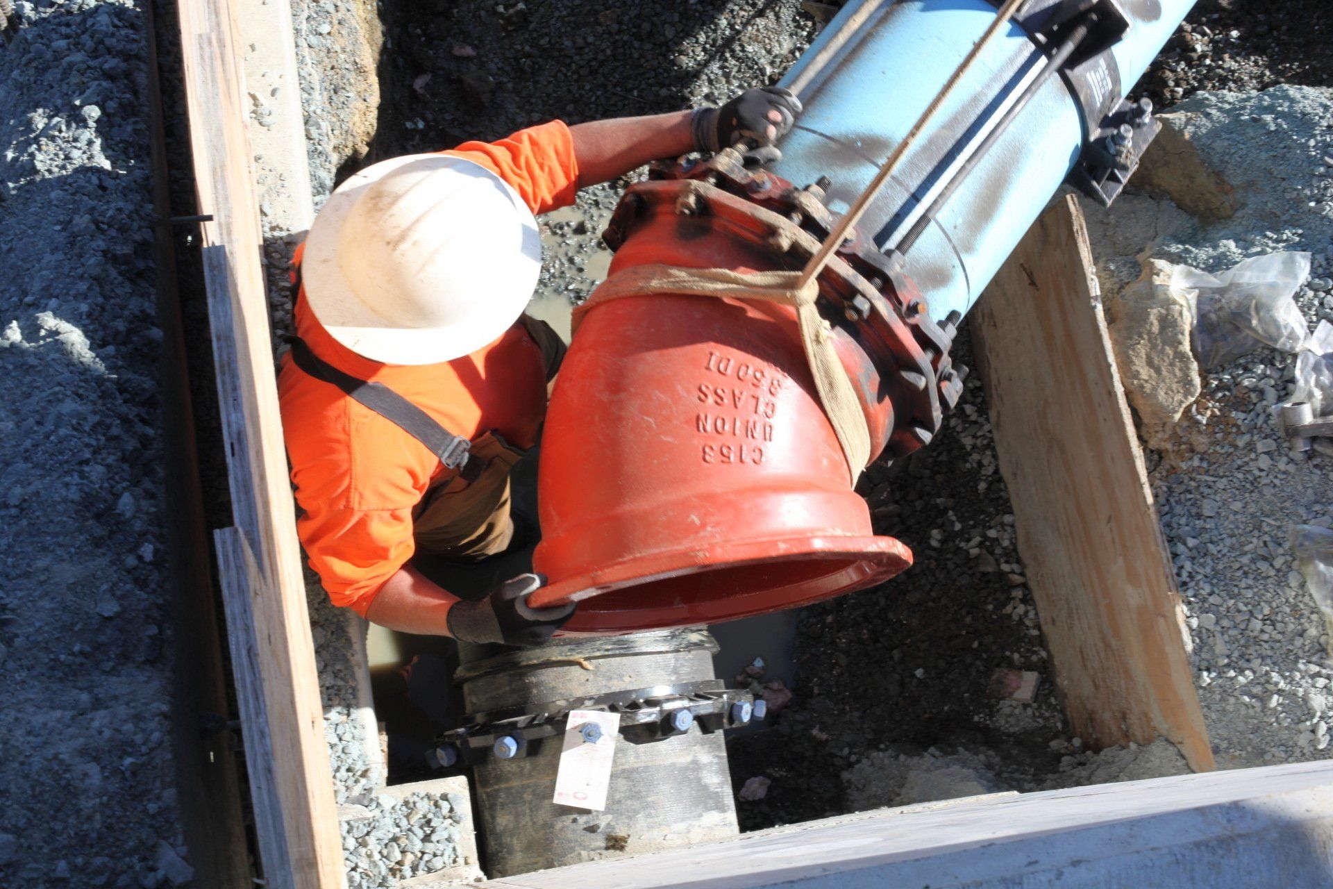 Construction worker connecting large pipes in a ditch, wearing orange shirt and white hard hat.