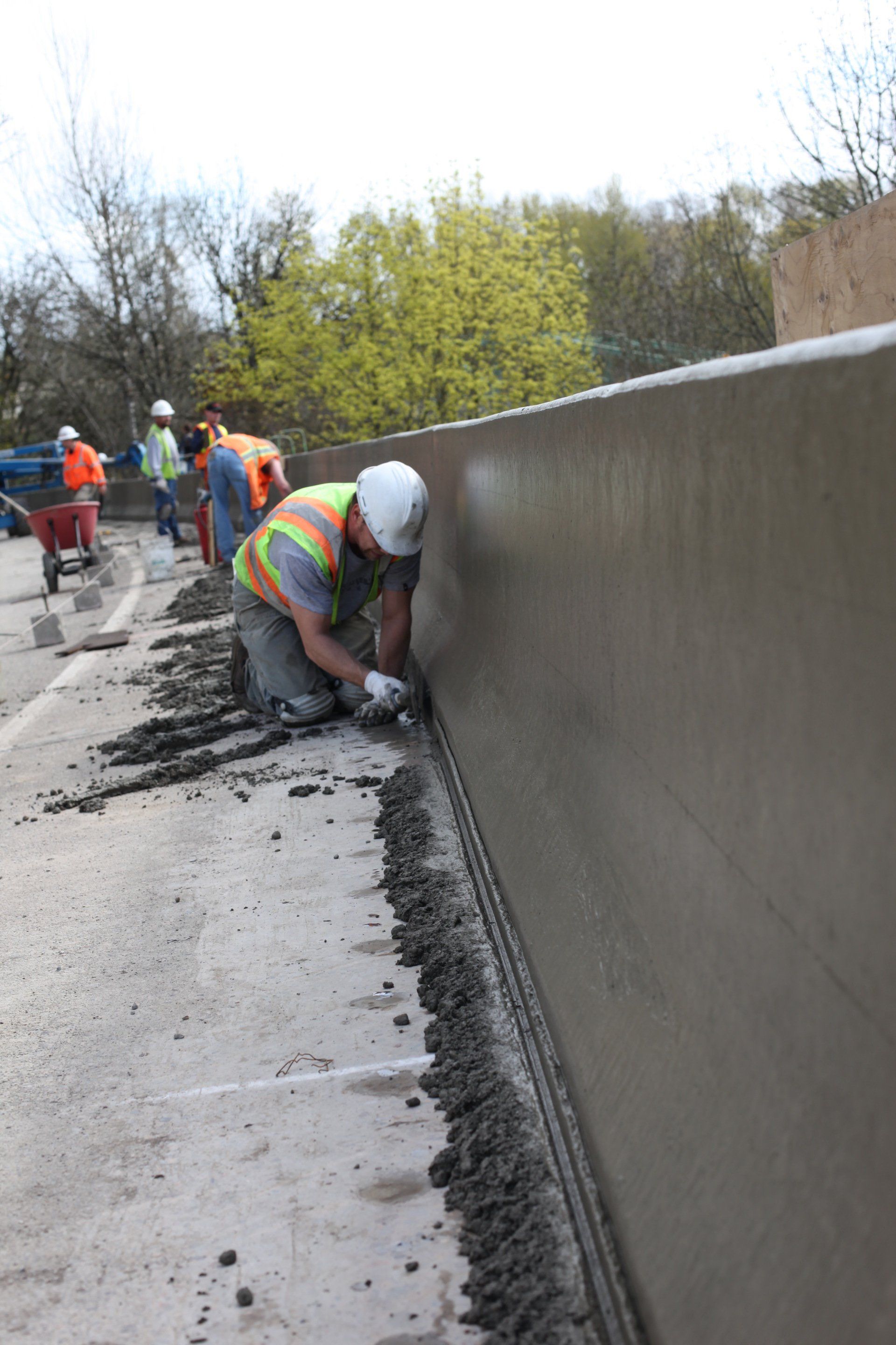 Construction worker kneels, using a trowel to smooth wet concrete on a roadside barrier.
