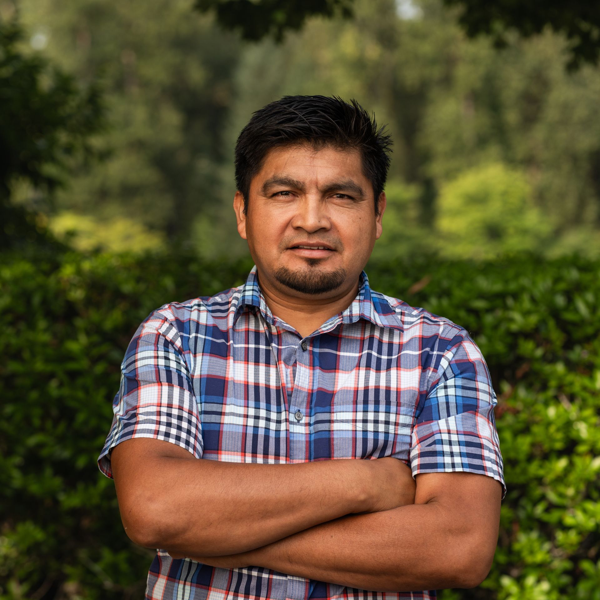 Man with arms crossed, wearing a blue plaid shirt, standing outdoors in front of green foliage.