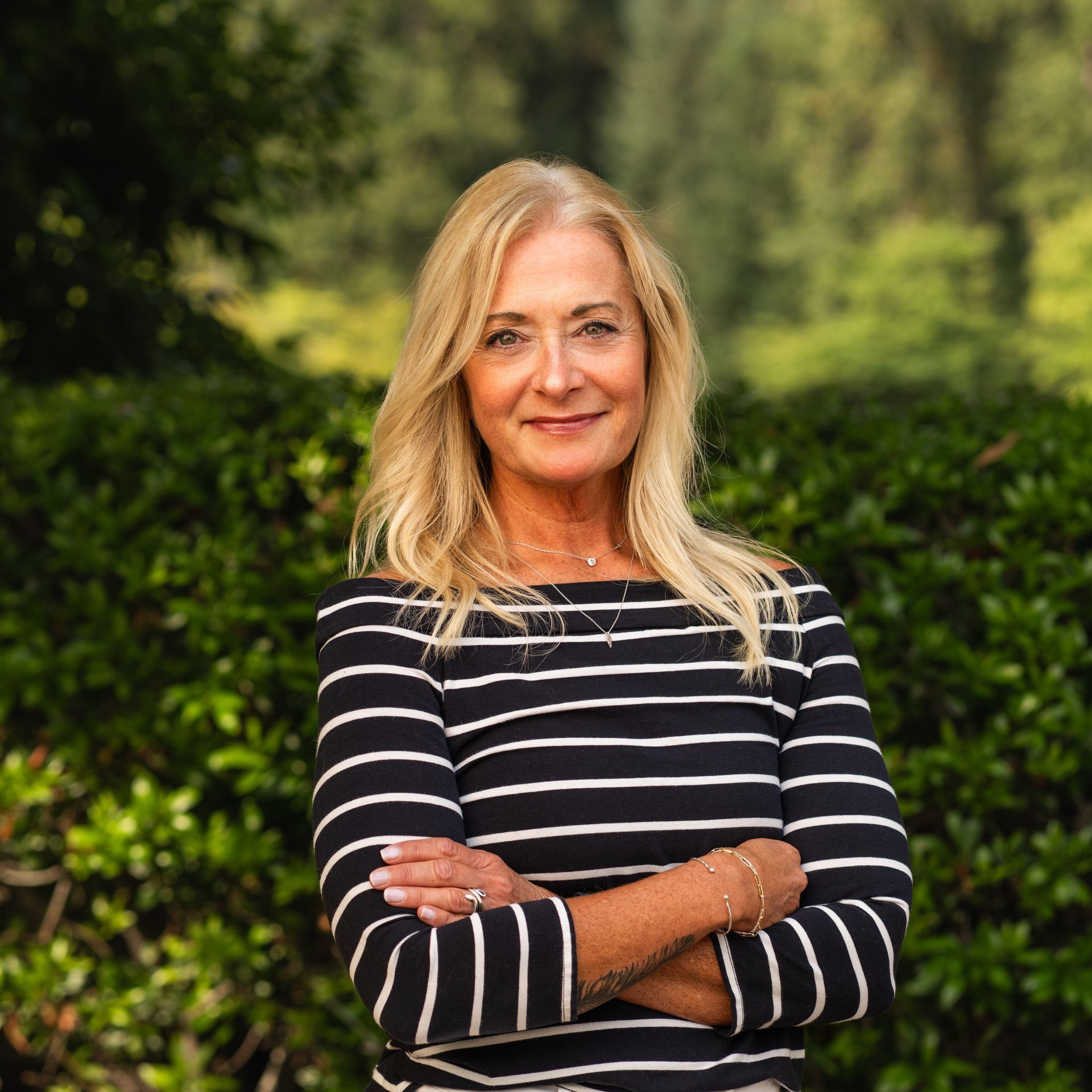 Woman with blonde hair, arms crossed, wearing a striped shirt outdoors near green foliage.