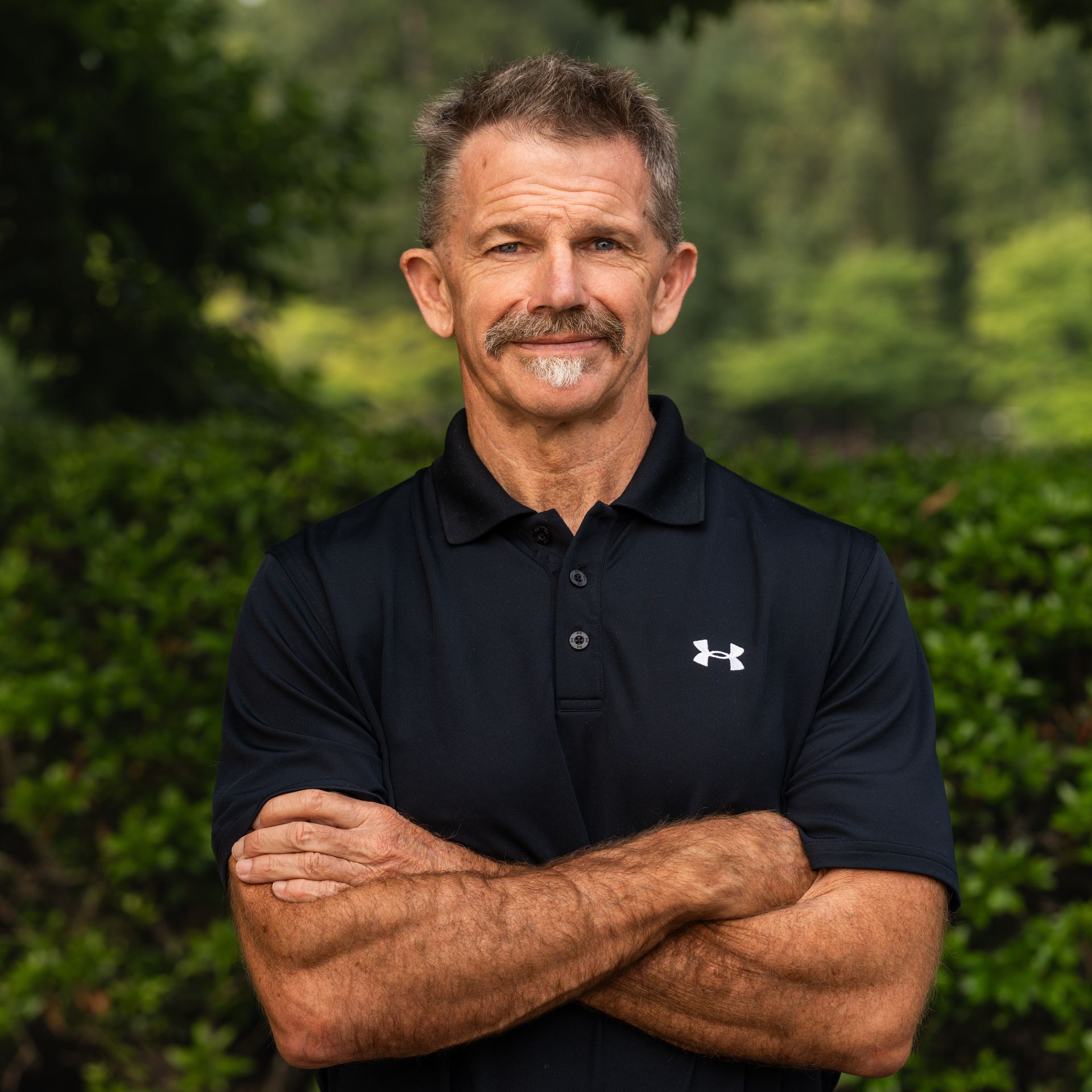 Man with crossed arms, wearing black polo shirt, standing outdoors with a green background.