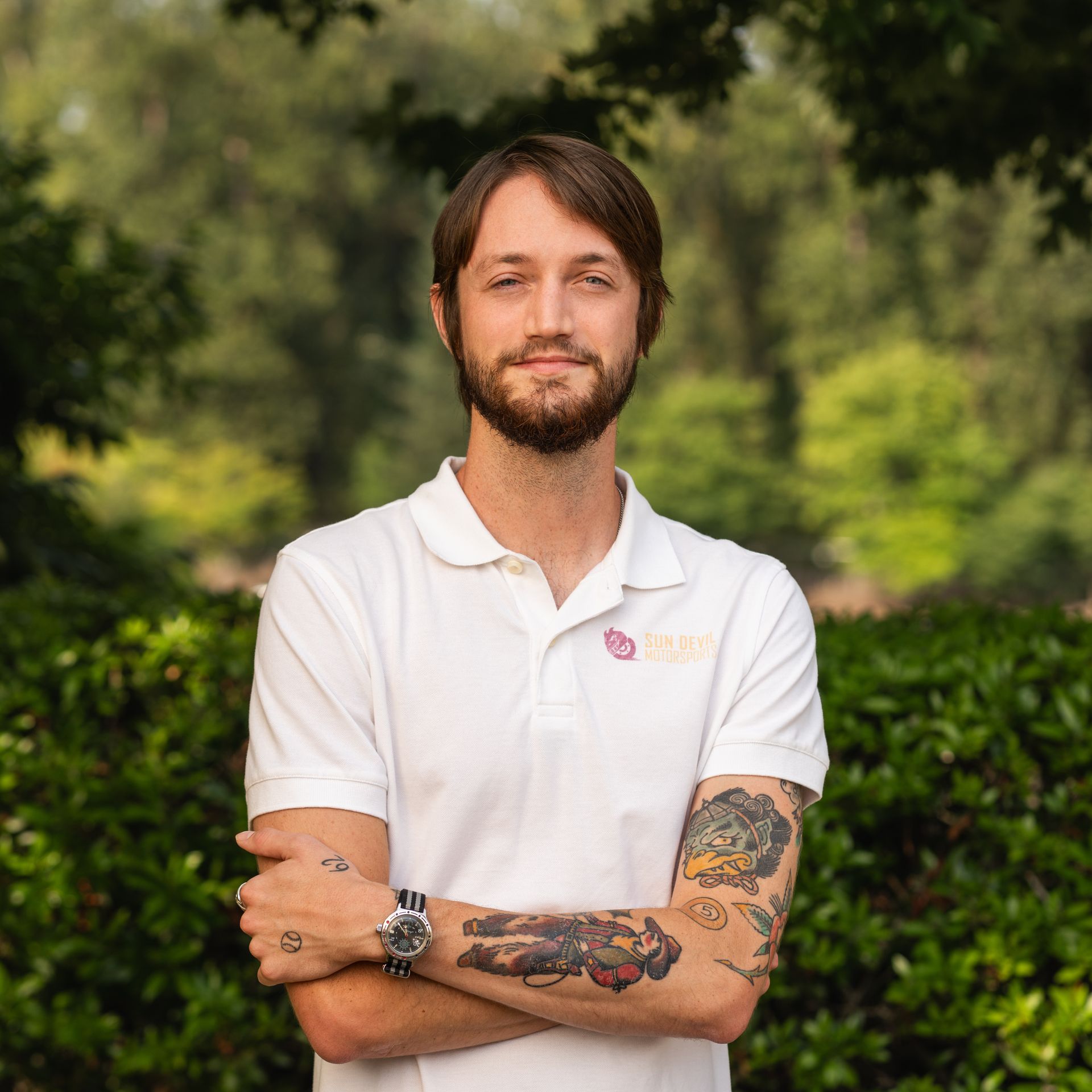 Man with tattoos wearing a white polo shirt, arms crossed, standing outdoors with green foliage in the background.