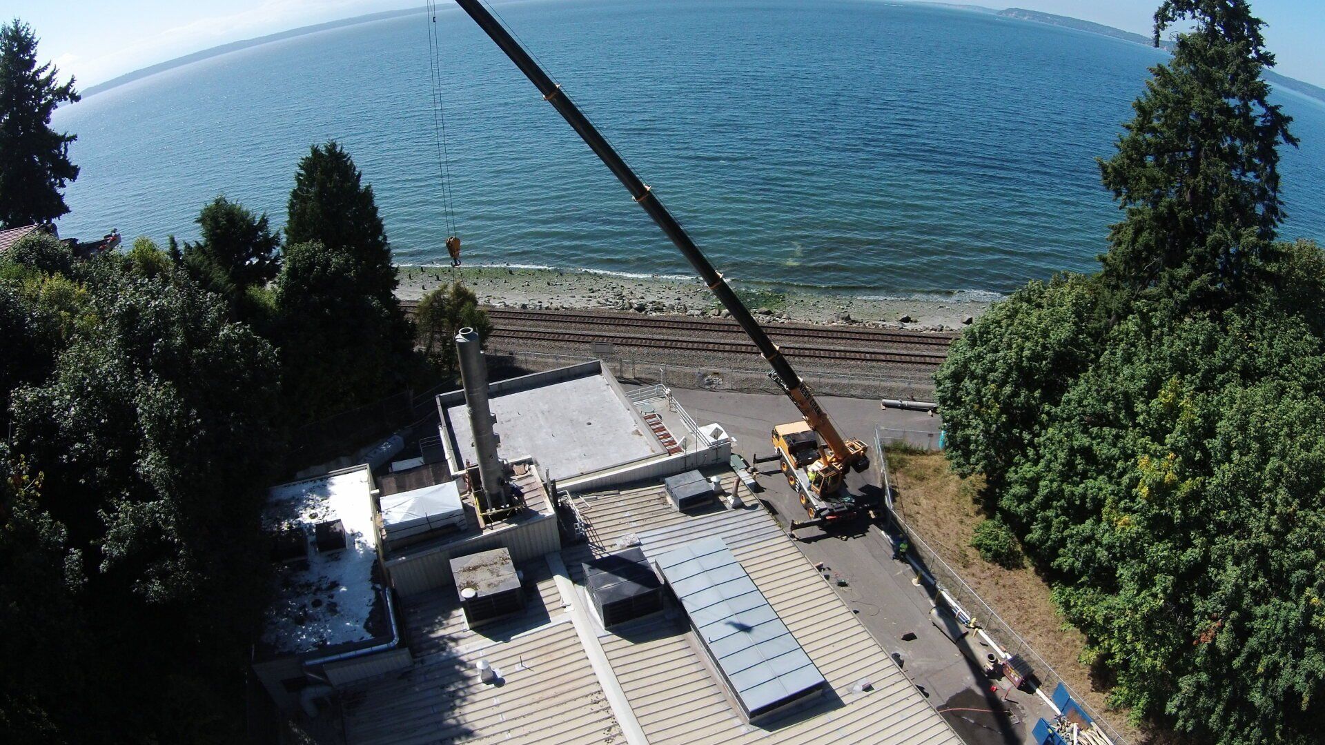Construction site near a body of water with crane, concrete structure, and trees. Blue water and sky.