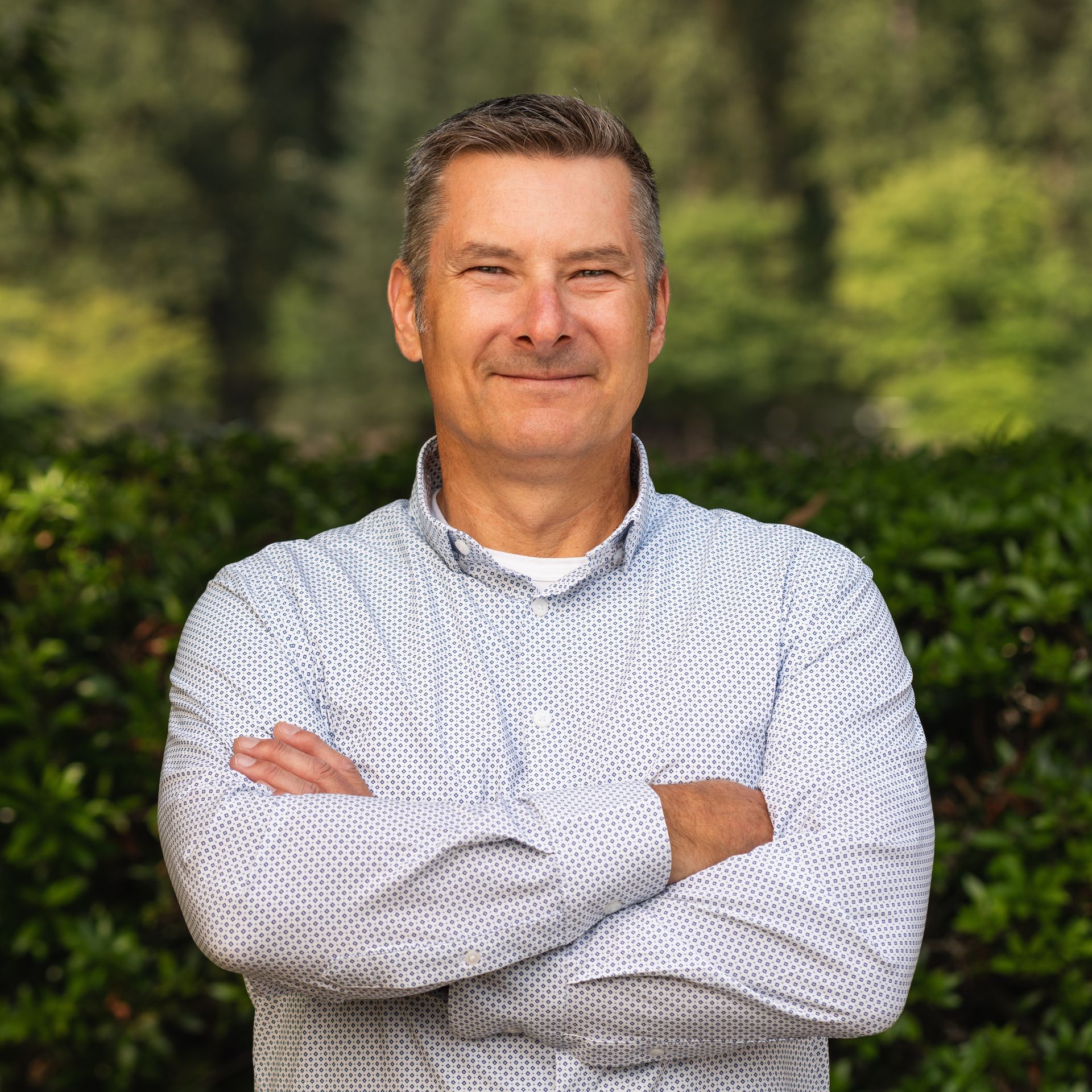 Man with arms crossed, smiling, wearing a light blue patterned shirt, set in front of green foliage.
