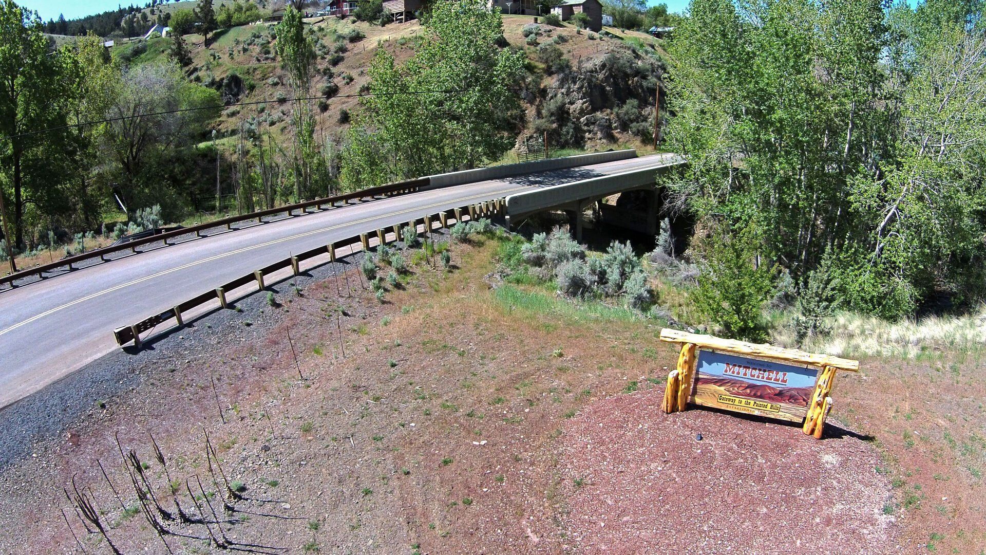 Bridge over a ravine; a wooden sign with artwork sits on a hill. Sunny, daytime.
