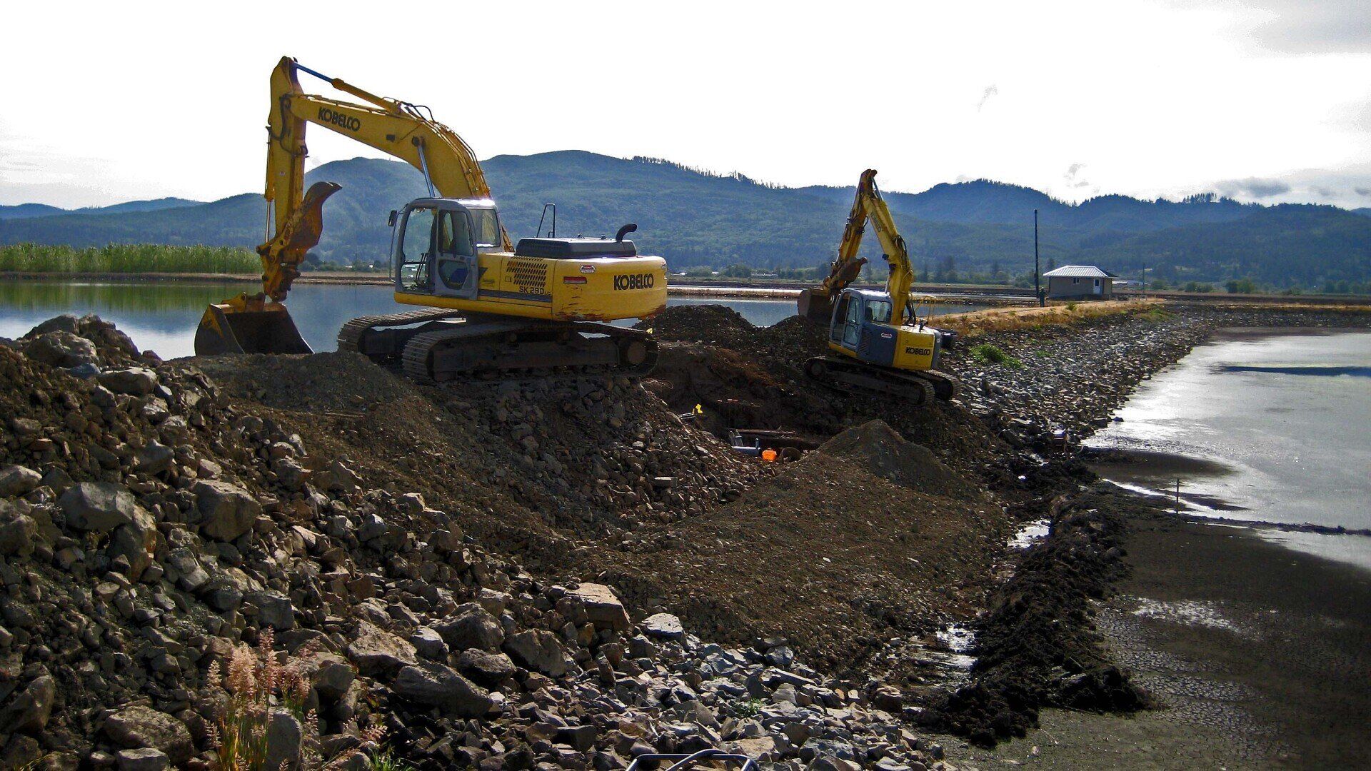 Two yellow excavators digging at a shoreline construction site; mountains in the background.