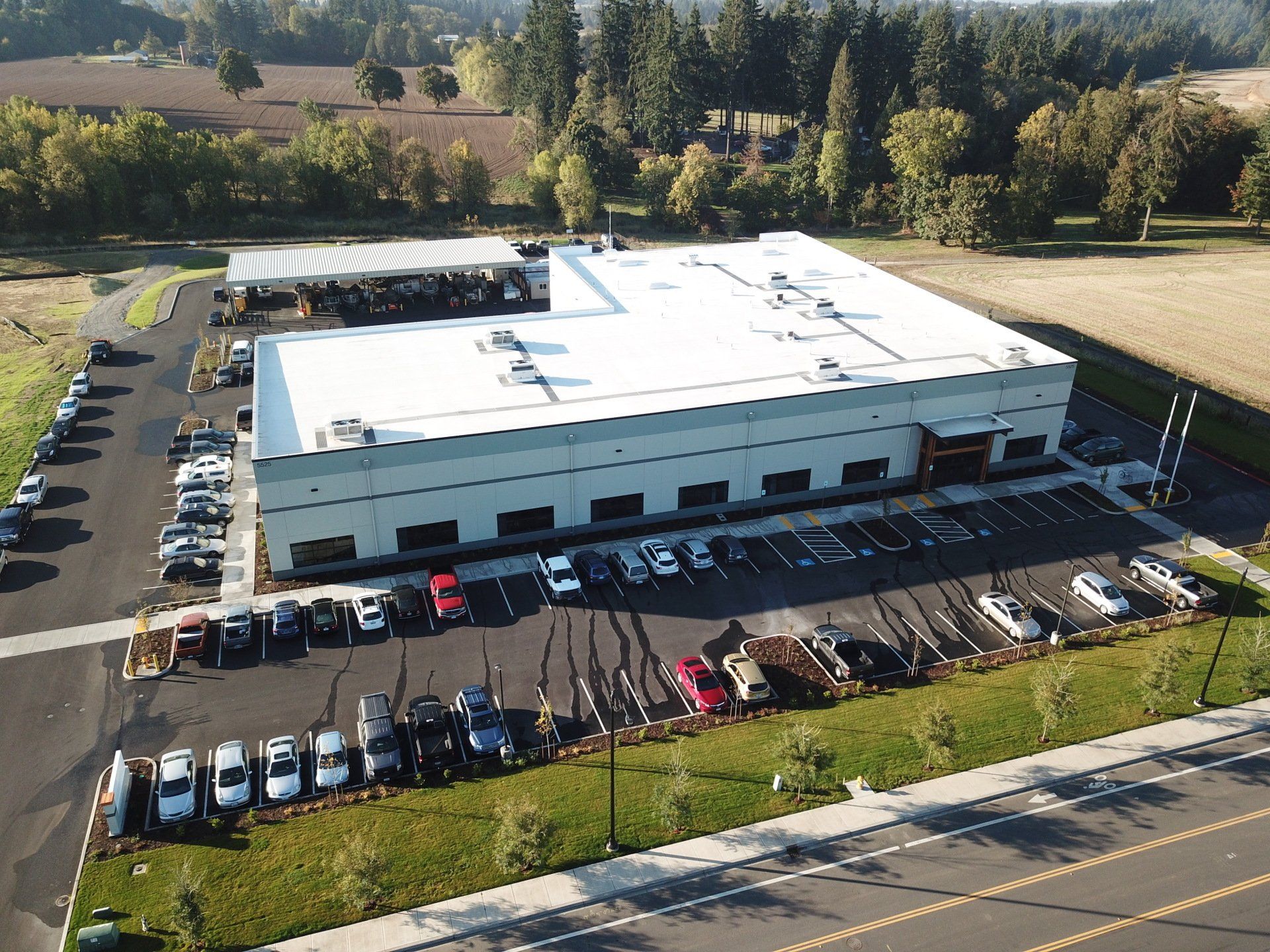 Large, light-colored industrial building with vehicles parked in front. Surrounding landscape includes grass and trees.