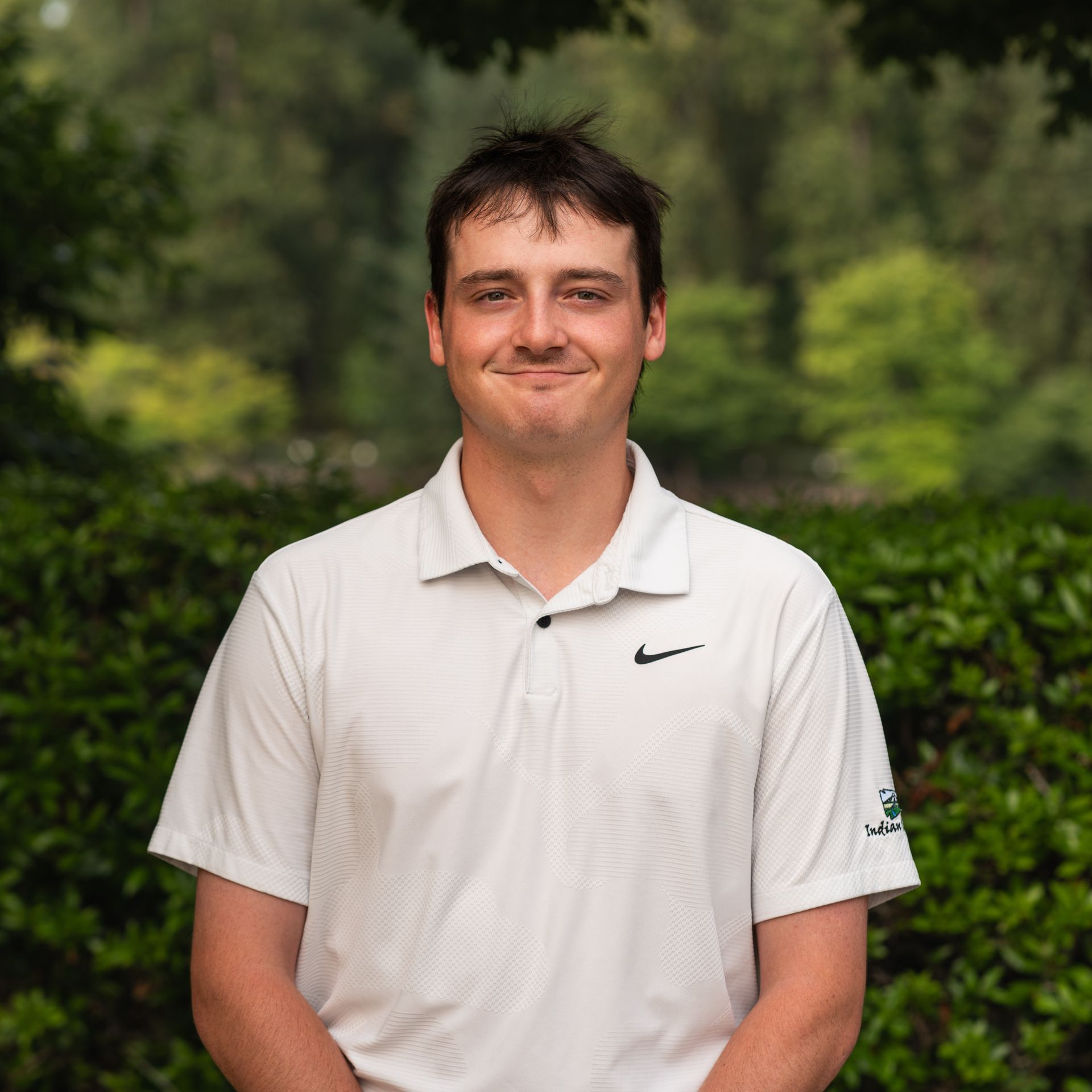 Man in white polo shirt smiles in front of green foliage.