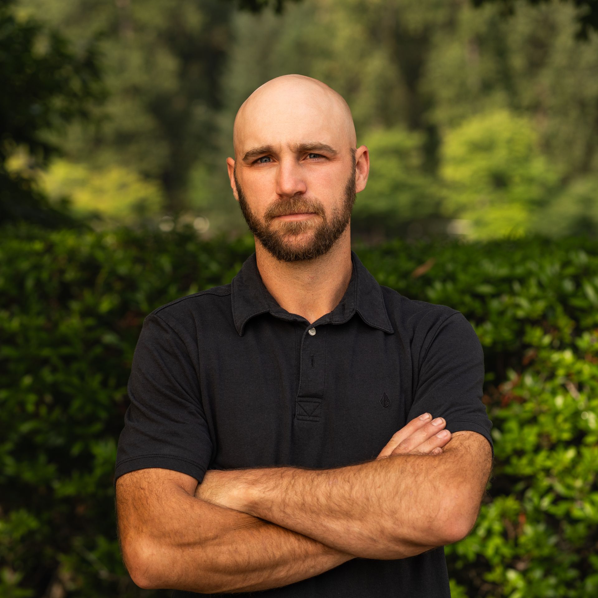 Bald man with beard and crossed arms, wearing a black shirt, set against a green, leafy background.