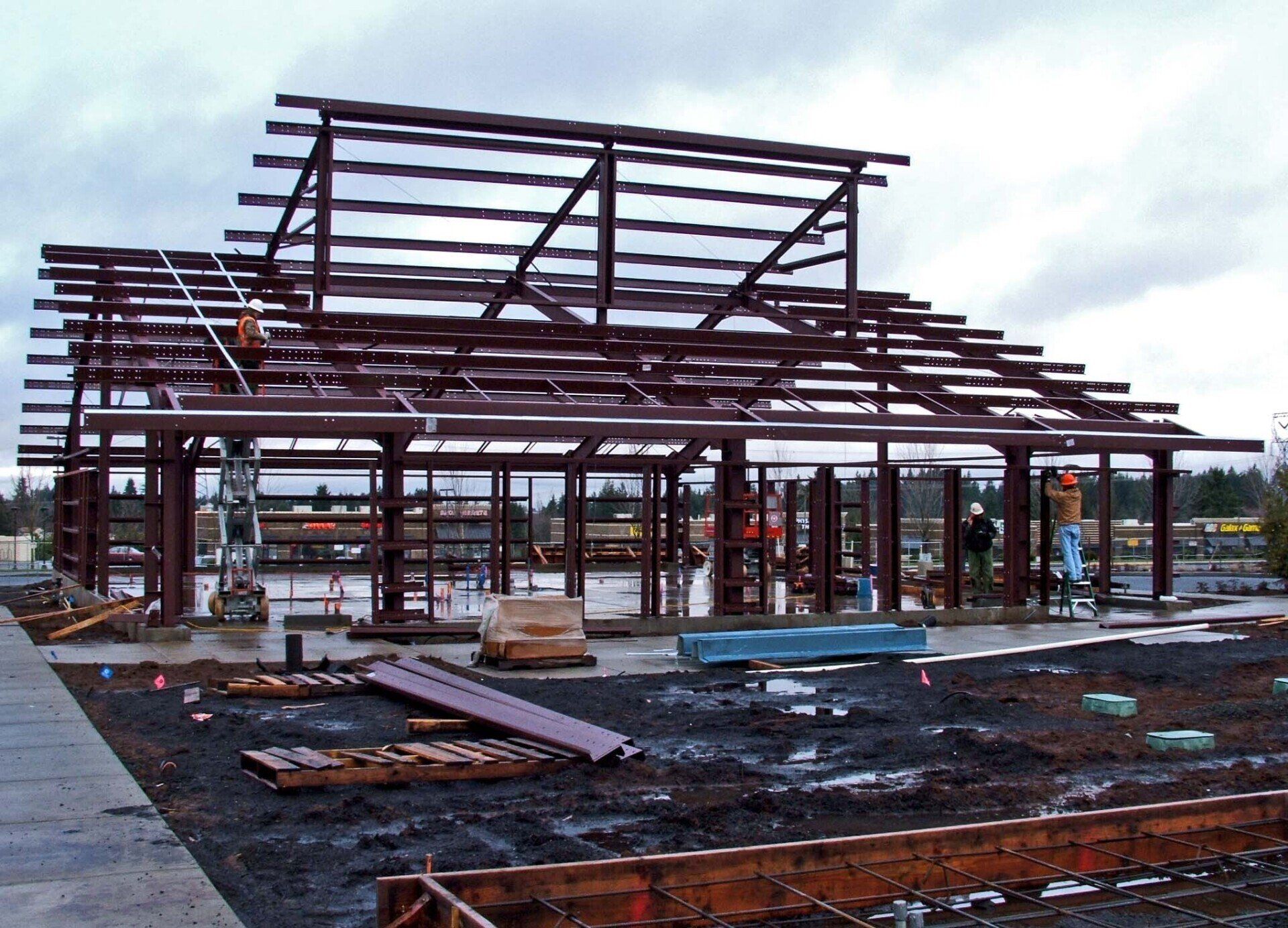 Steel frame of a building under construction, brown against a cloudy sky.