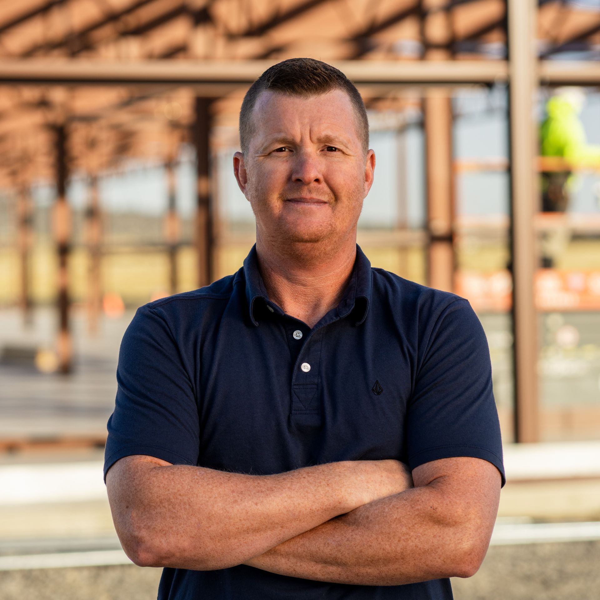 Man in navy polo shirt with arms crossed, standing outdoors in front of a building.