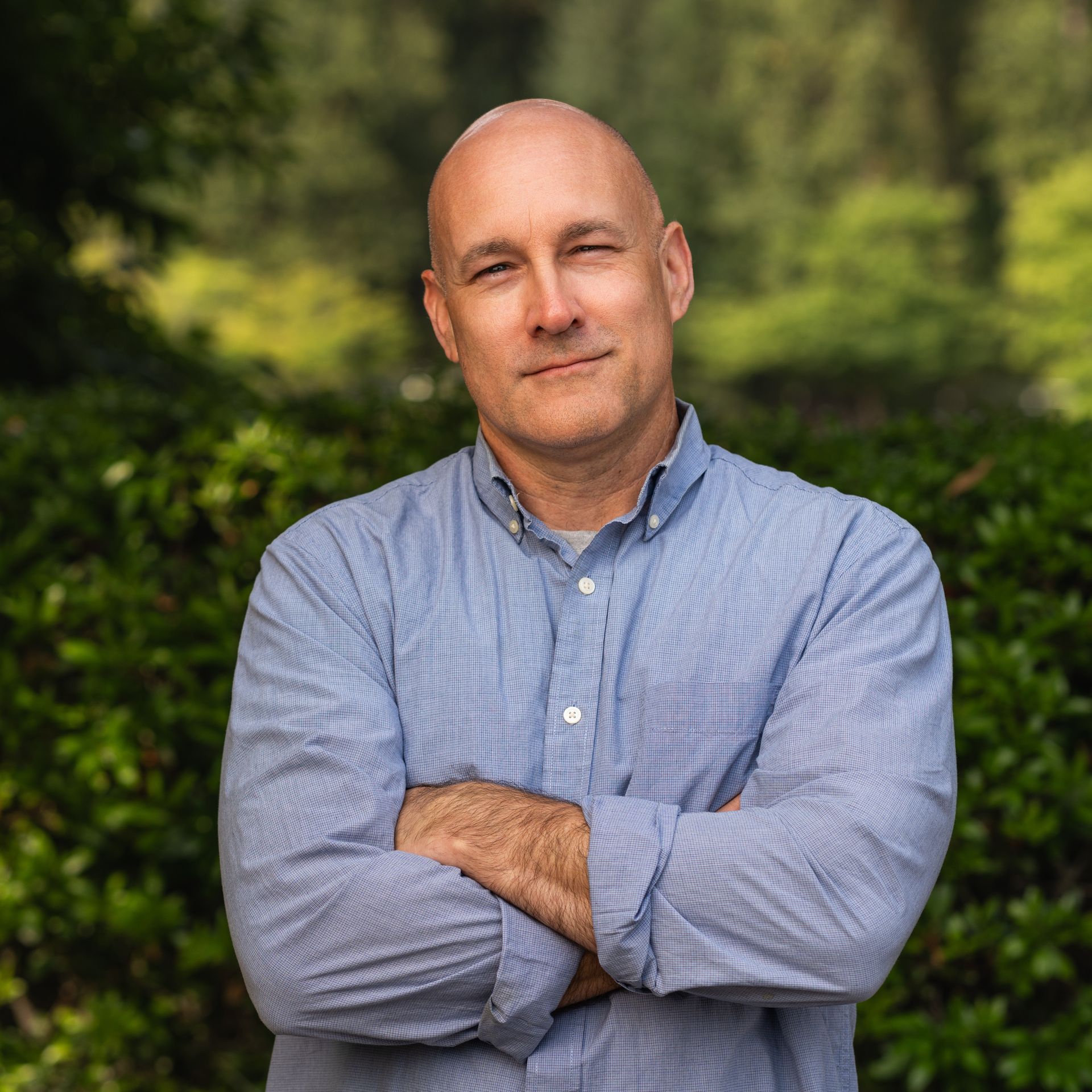 Bald man in blue button-down shirt with arms crossed, smiling slightly, standing outside near green foliage.