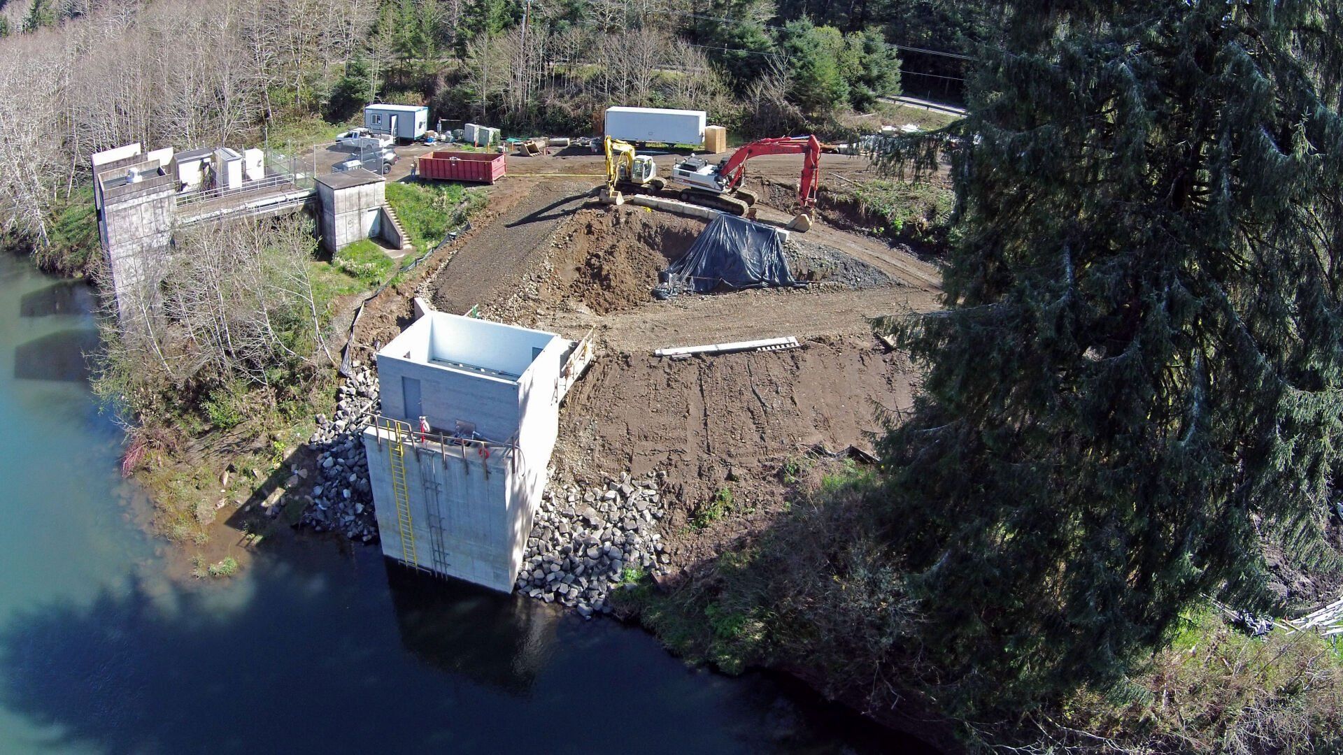 Construction site on a riverbank with a partially demolished concrete structure. An excavator is present with dirt and debris.