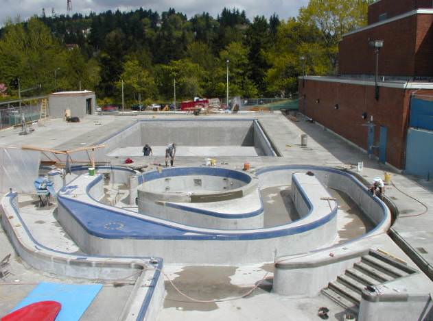 Pool under construction: Concrete basins, partially tiled, with workers, surrounded by concrete deck and building.