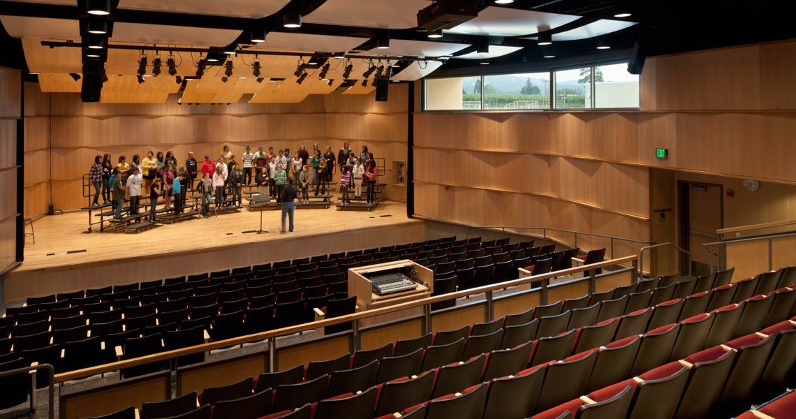 Auditorium with choir on stage, audience seating, wooden paneling, and a window.