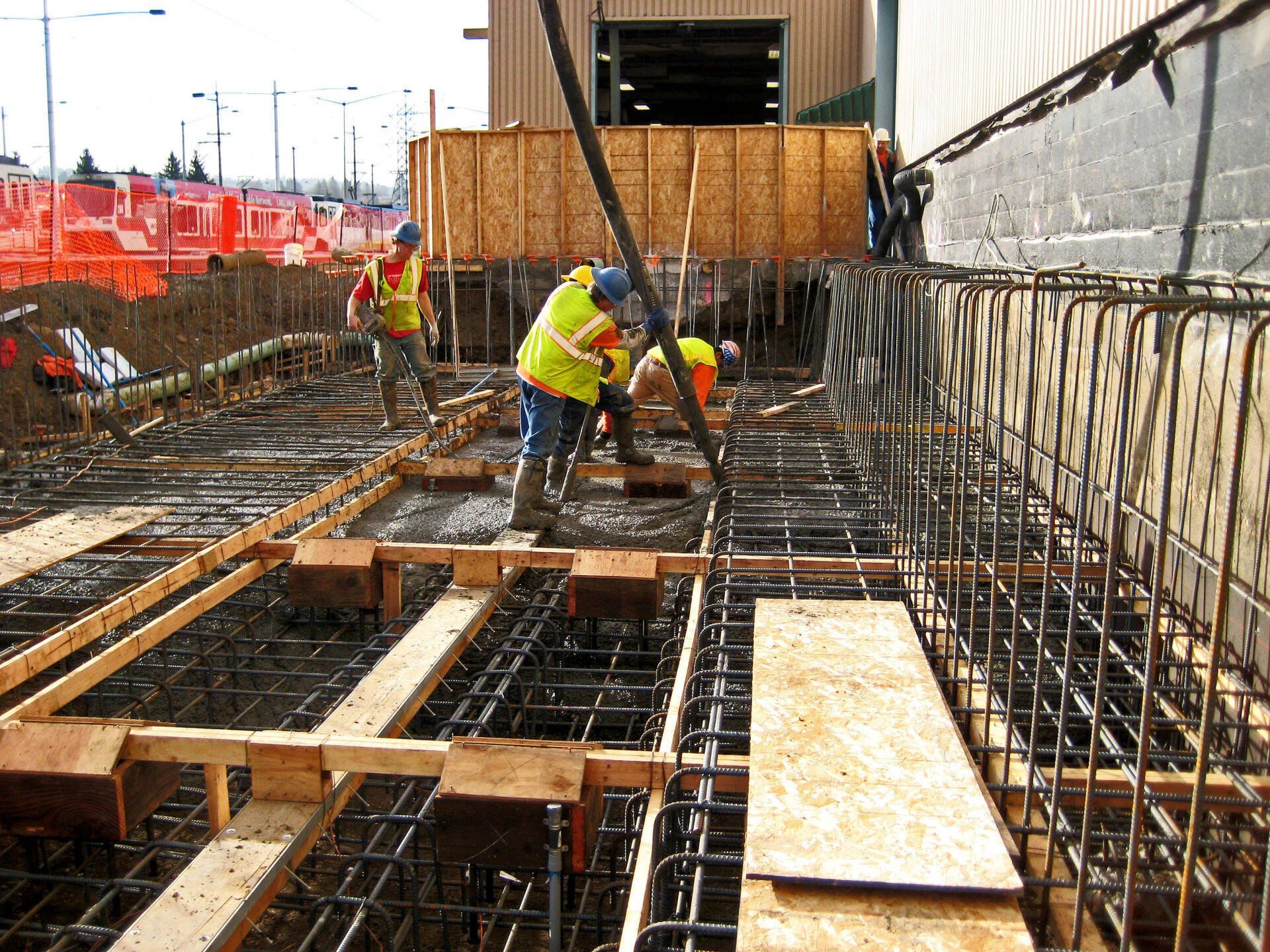 Construction workers pouring concrete at a construction site. Steel rebar and wooden forms visible.