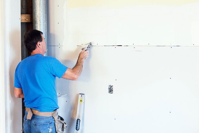 Man in blue shirt, applying drywall compound to wall with a trowel.