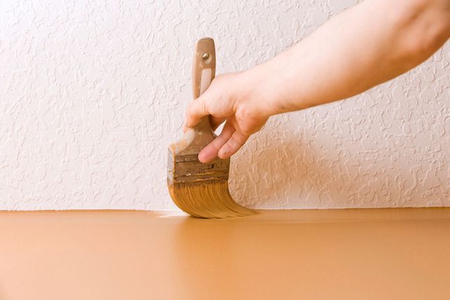 Hand painting a smooth, light brown surface with a brush, against a textured white wall.