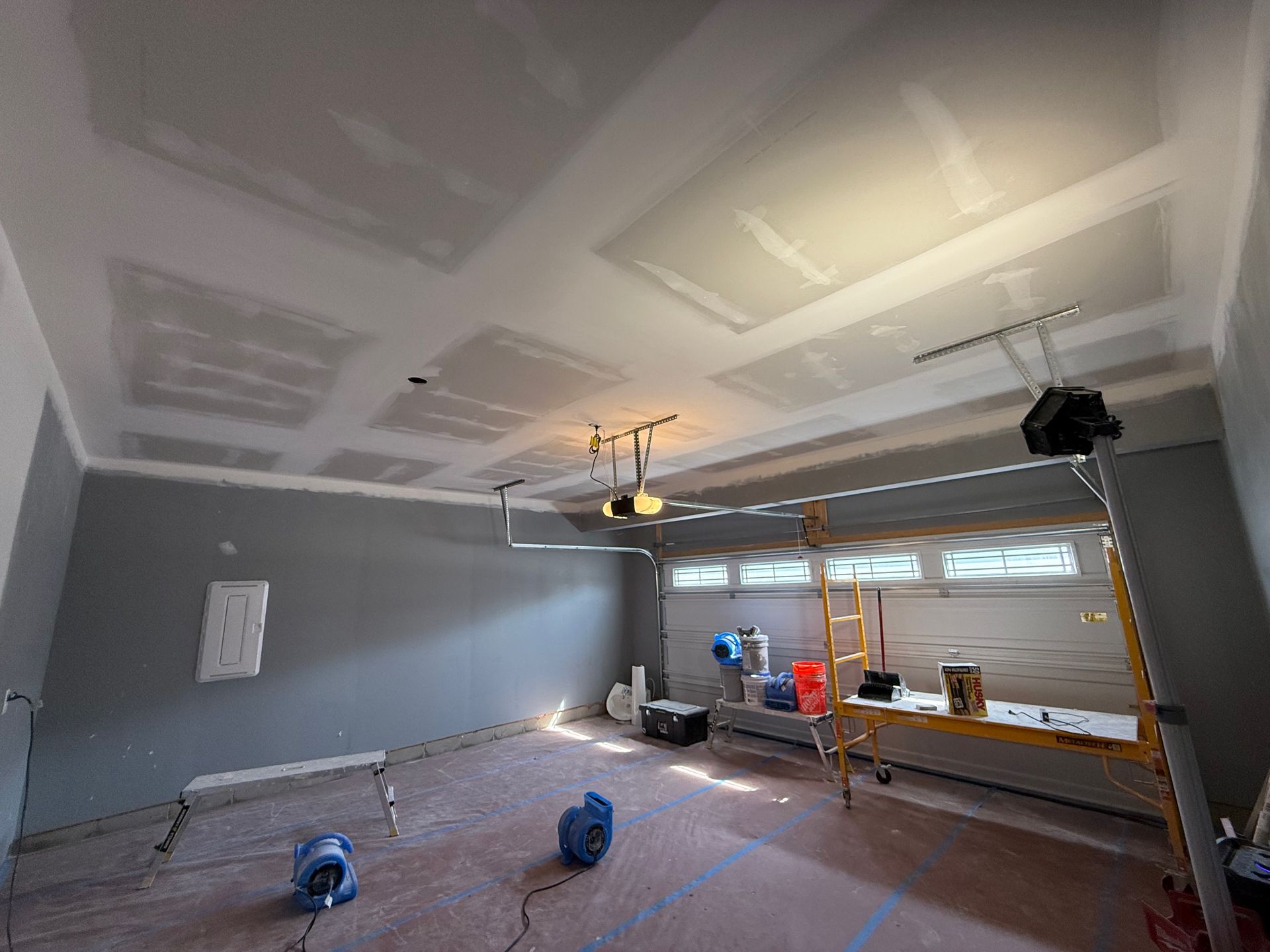 Garage interior under renovation. Gray walls, white ceiling with recessed panels, tools and equipment present.