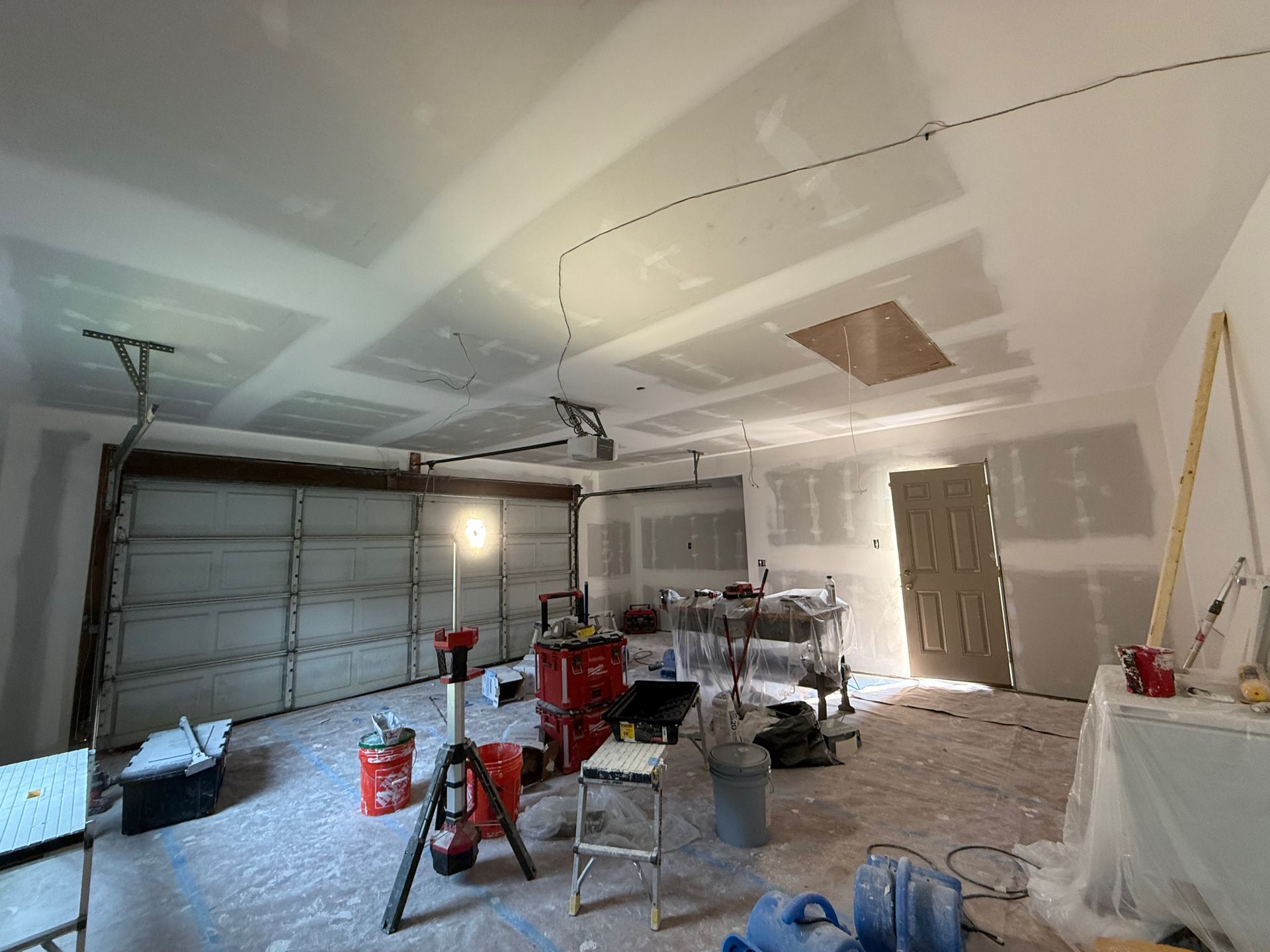 Garage interior under construction: drywall installed on ceiling and walls, tools and equipment scattered about.