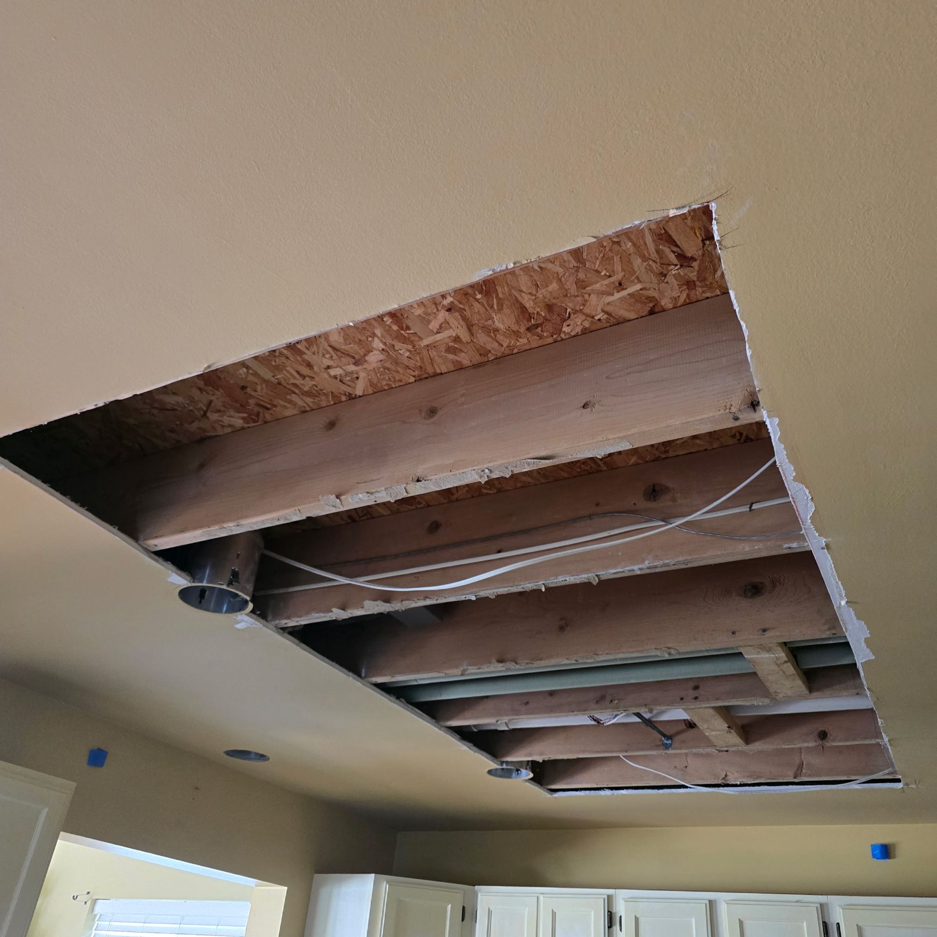 Hole cut in a yellow ceiling, exposing wooden joists, pipes, and electrical wiring.