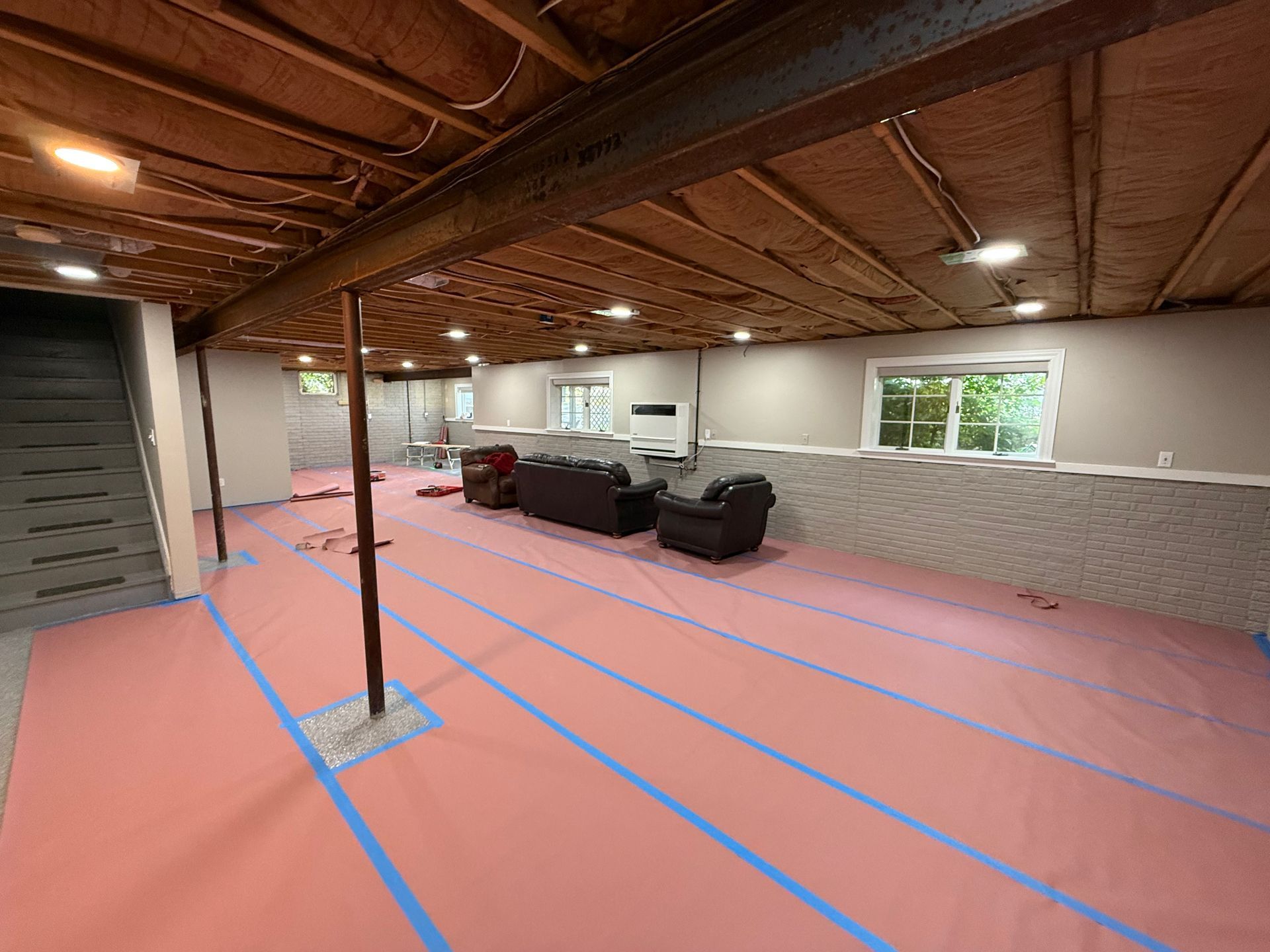 Basement renovation: Pink flooring, blue tape lines, exposed ceiling, support beams, furniture covered in plastic.