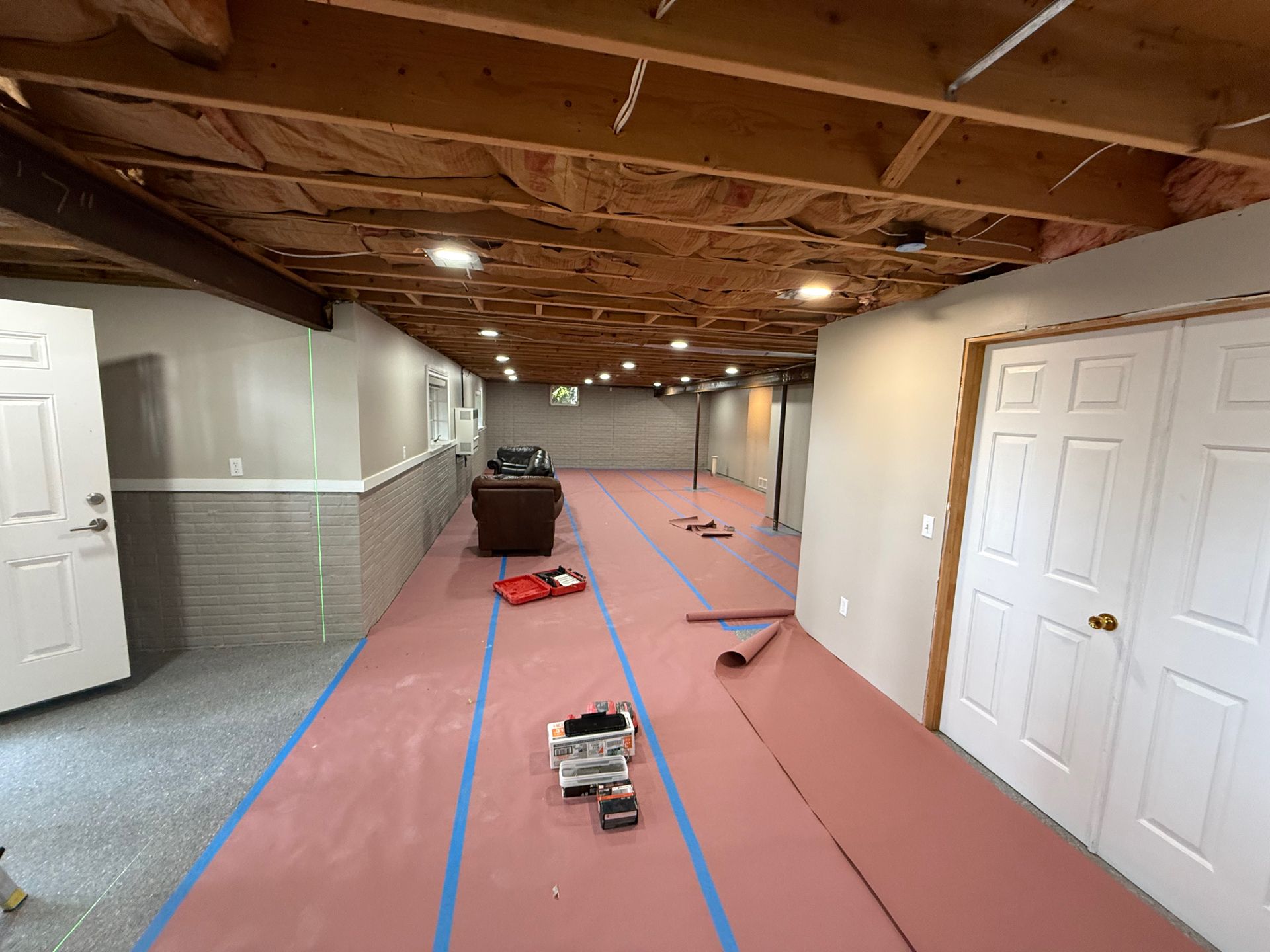 Basement renovation: Pink flooring, tools, and blue tape on the floor. Walls are gray/white, with a wood ceiling.