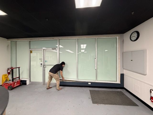 A person in a black shirt and tan pants uses a tape measure to measure the base of a glass-walled office front.