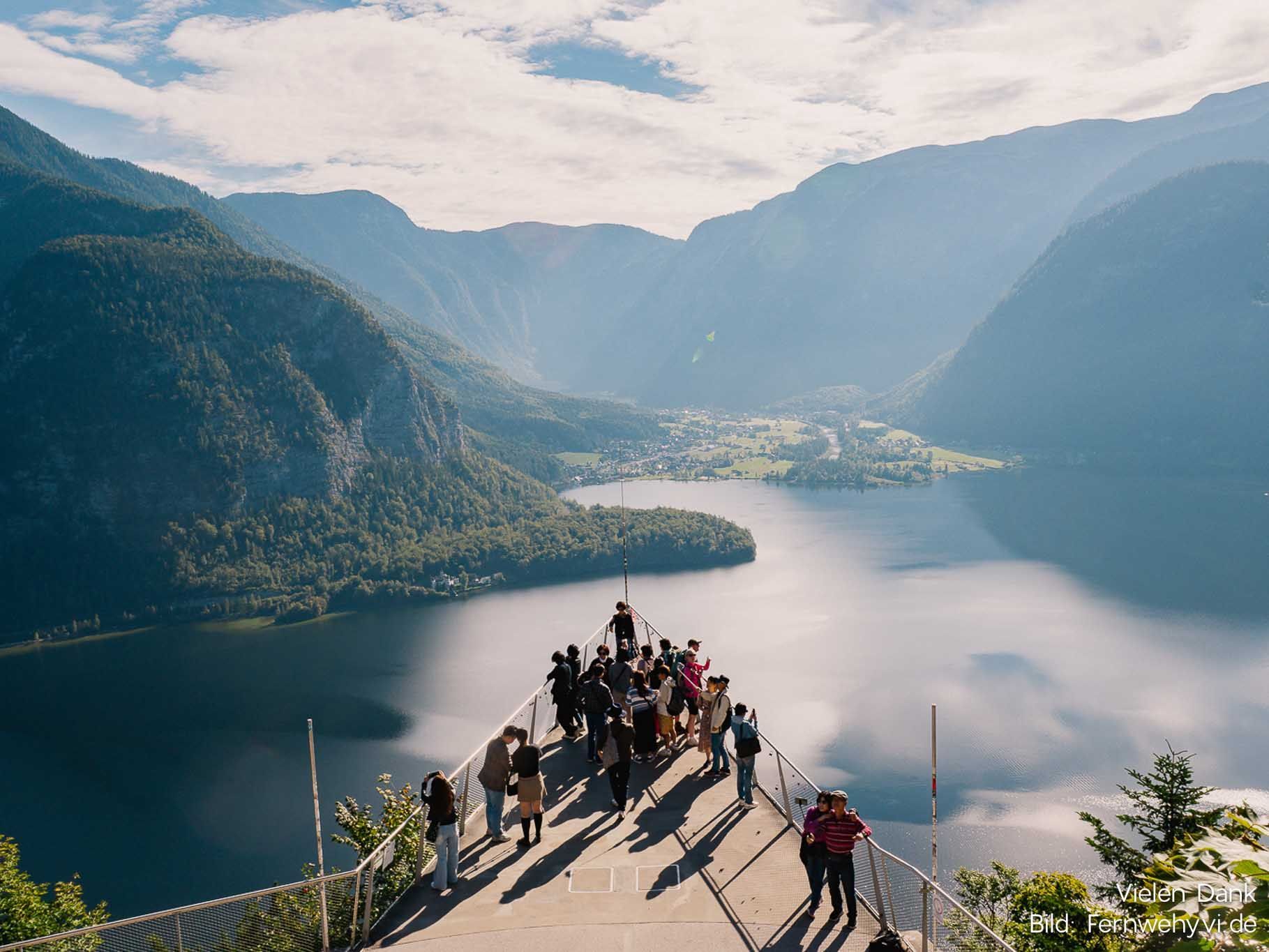 Menschen stehen auf einem Berggipfel mit Blick auf einen blauen See und ein Tal, in der Ferne erheben sich bewaldete Gipfel.