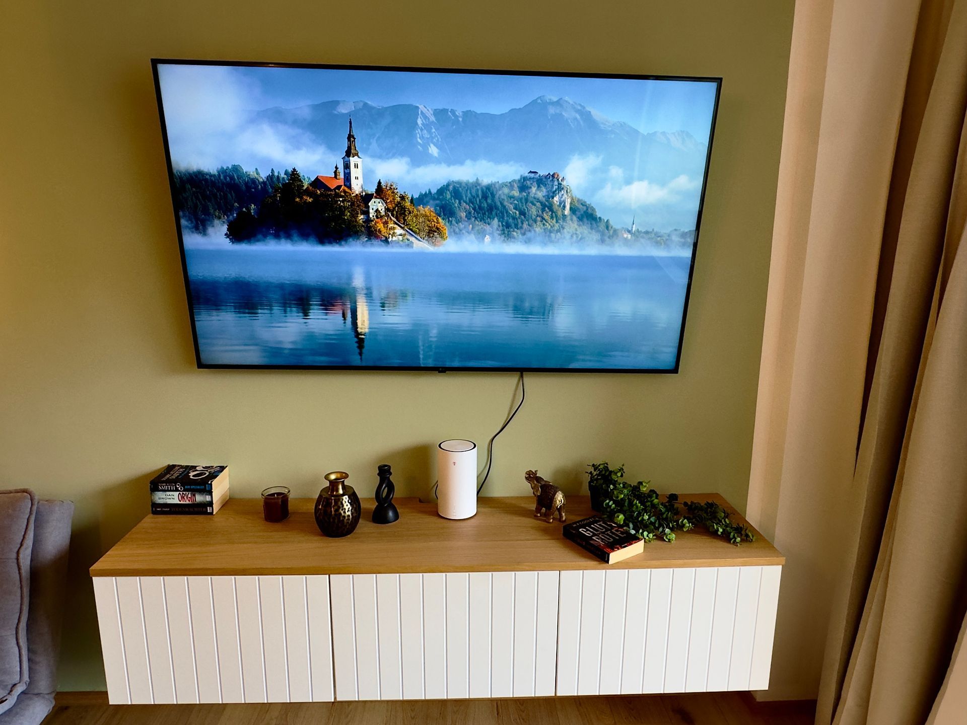 In einem beigen Zimmer hängt ein an der Wand montierter Fernseher, der eine Kirchenszene am See zeigt