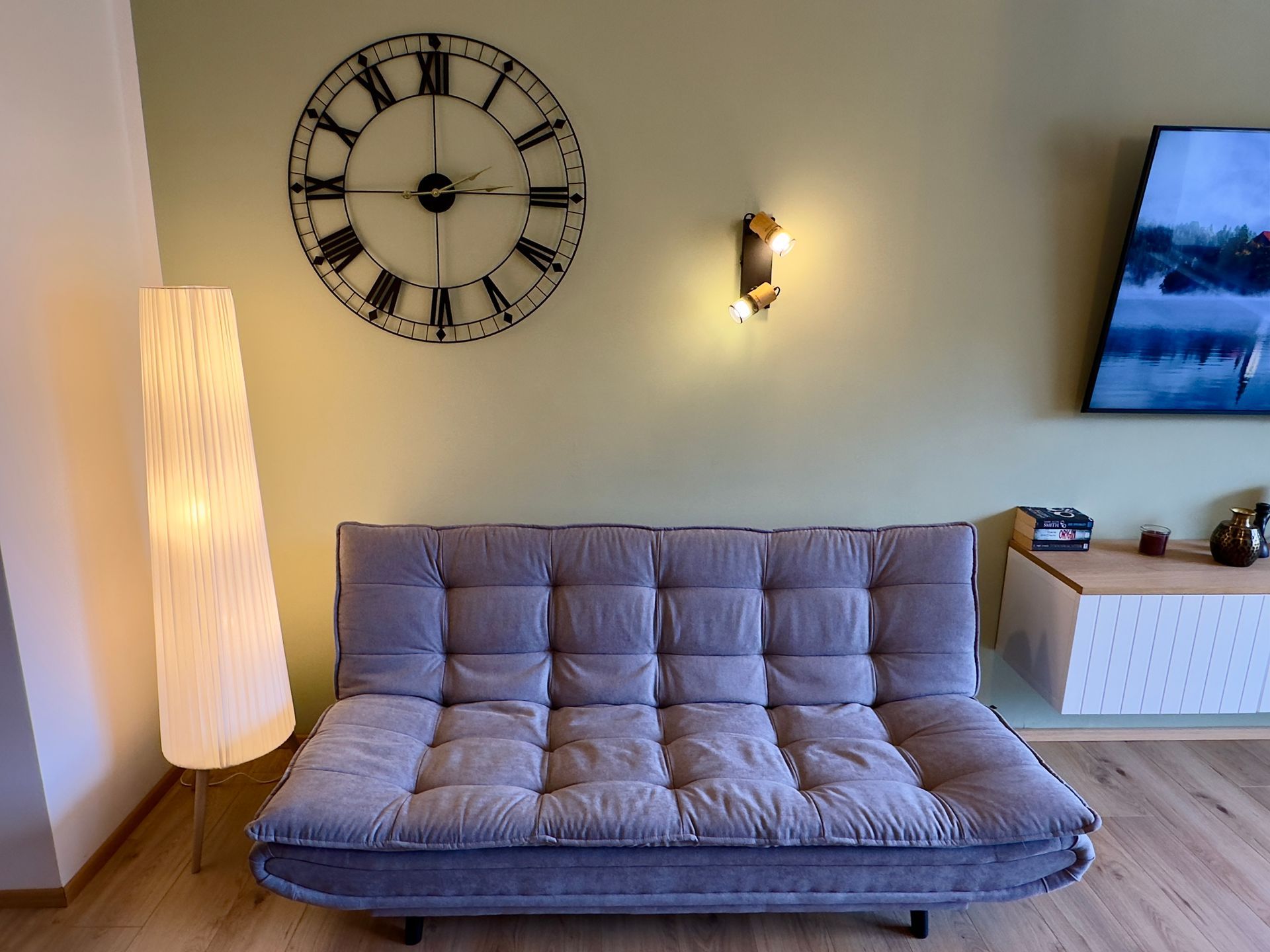 Minimalistisches Wohnzimmer mit grauem Sofa, großer Wanduhr, Stehlampe, Wandleuchte und Fernseher rechts.
