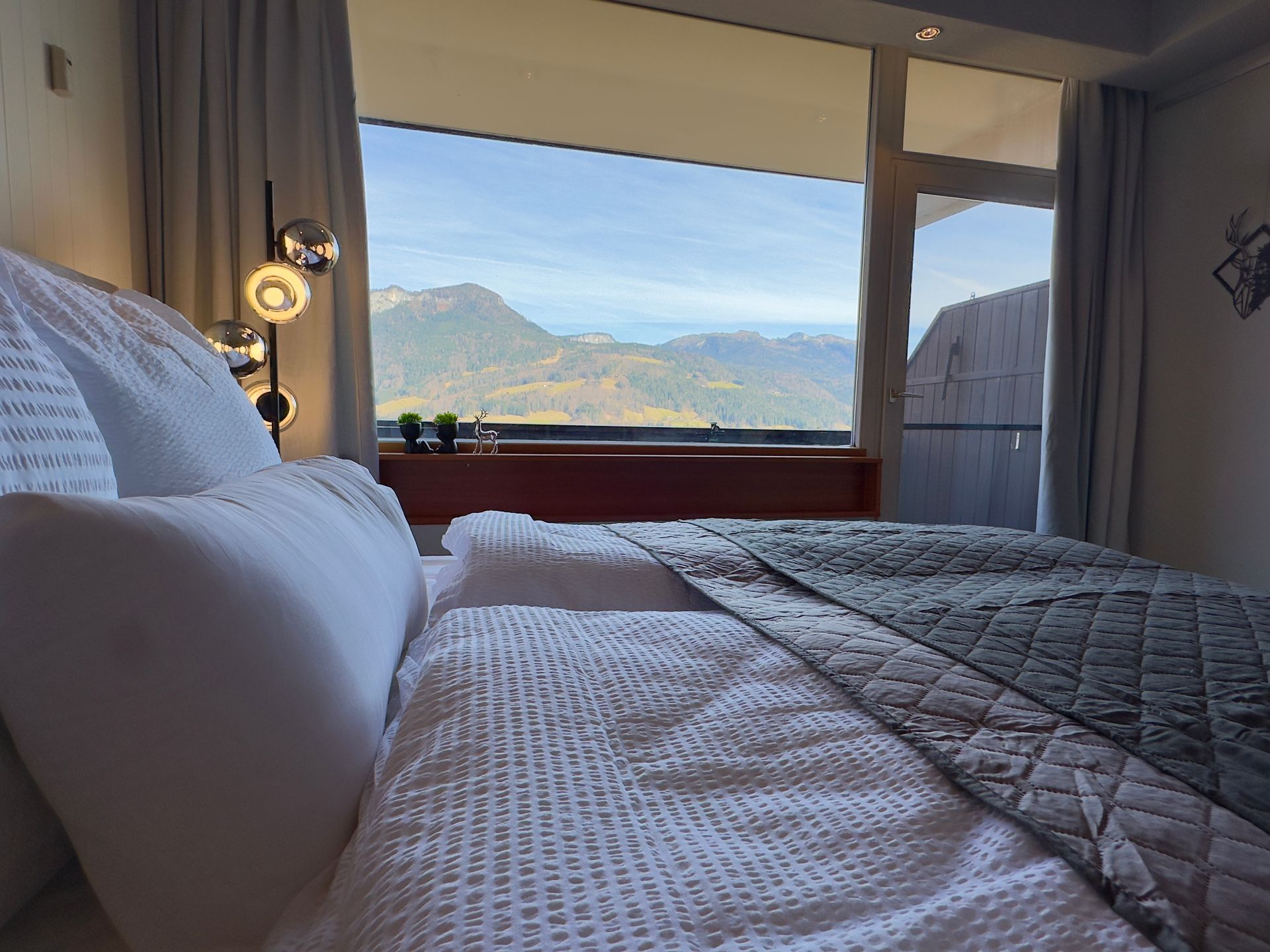 Gemütliches Bett mit Fensterblick auf Berge und Himmel, sanftes Lampenlicht in einem modernen Schlafzimmer