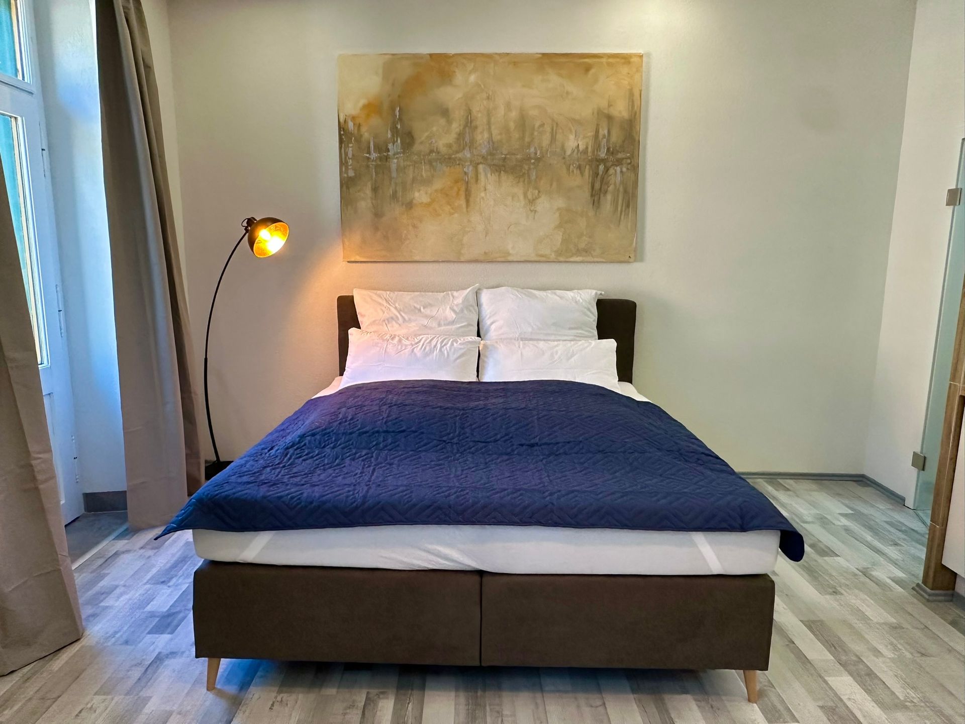 Minimalistisches Schlafzimmer mit blauer Tagesdecke, Stehlampe und abstrakter Wandkunst