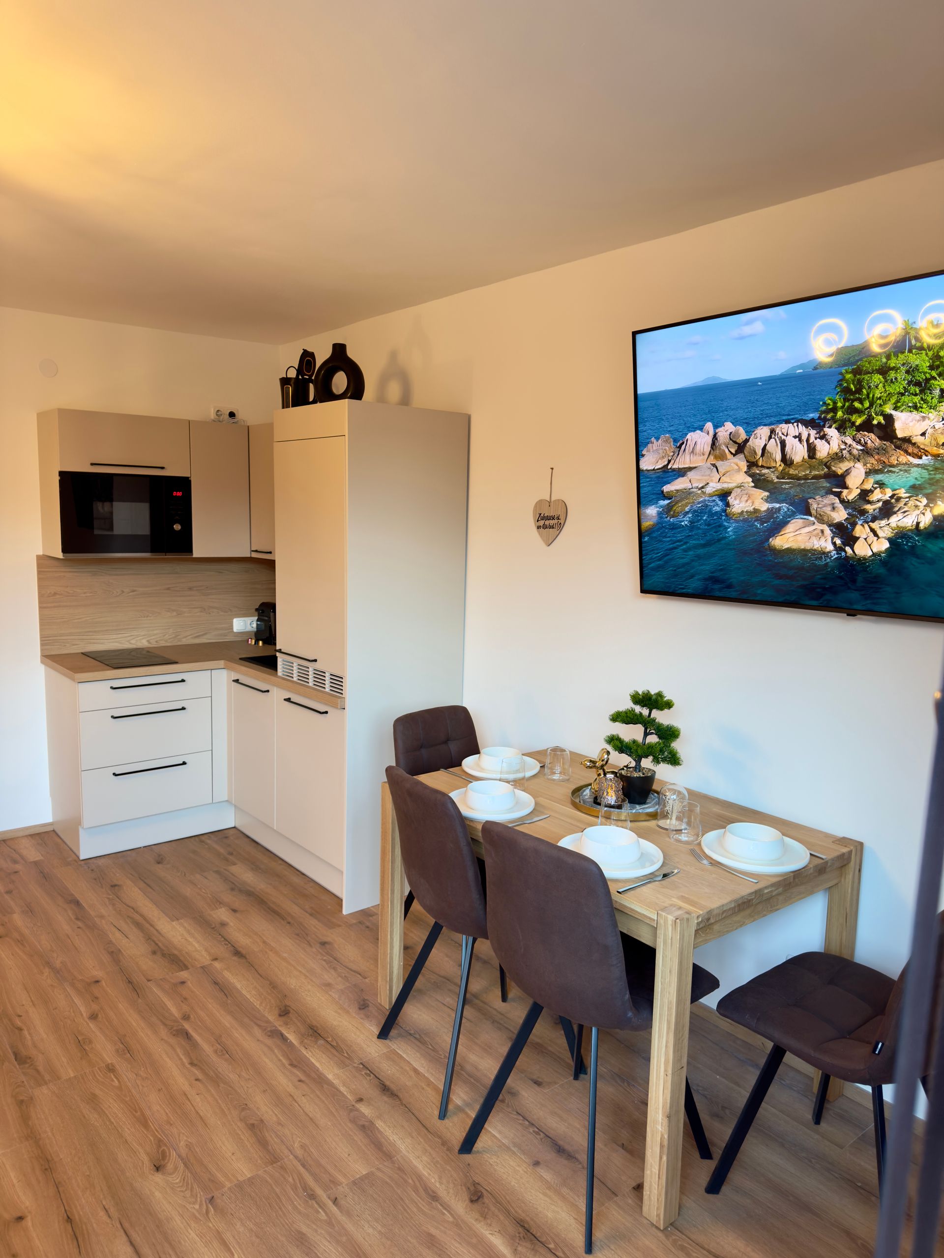 Kleine, moderne Küche mit Essbereich, Holzboden, weißen Schränken und einem Fernseher, der ein Küstenpanorama zeigt.