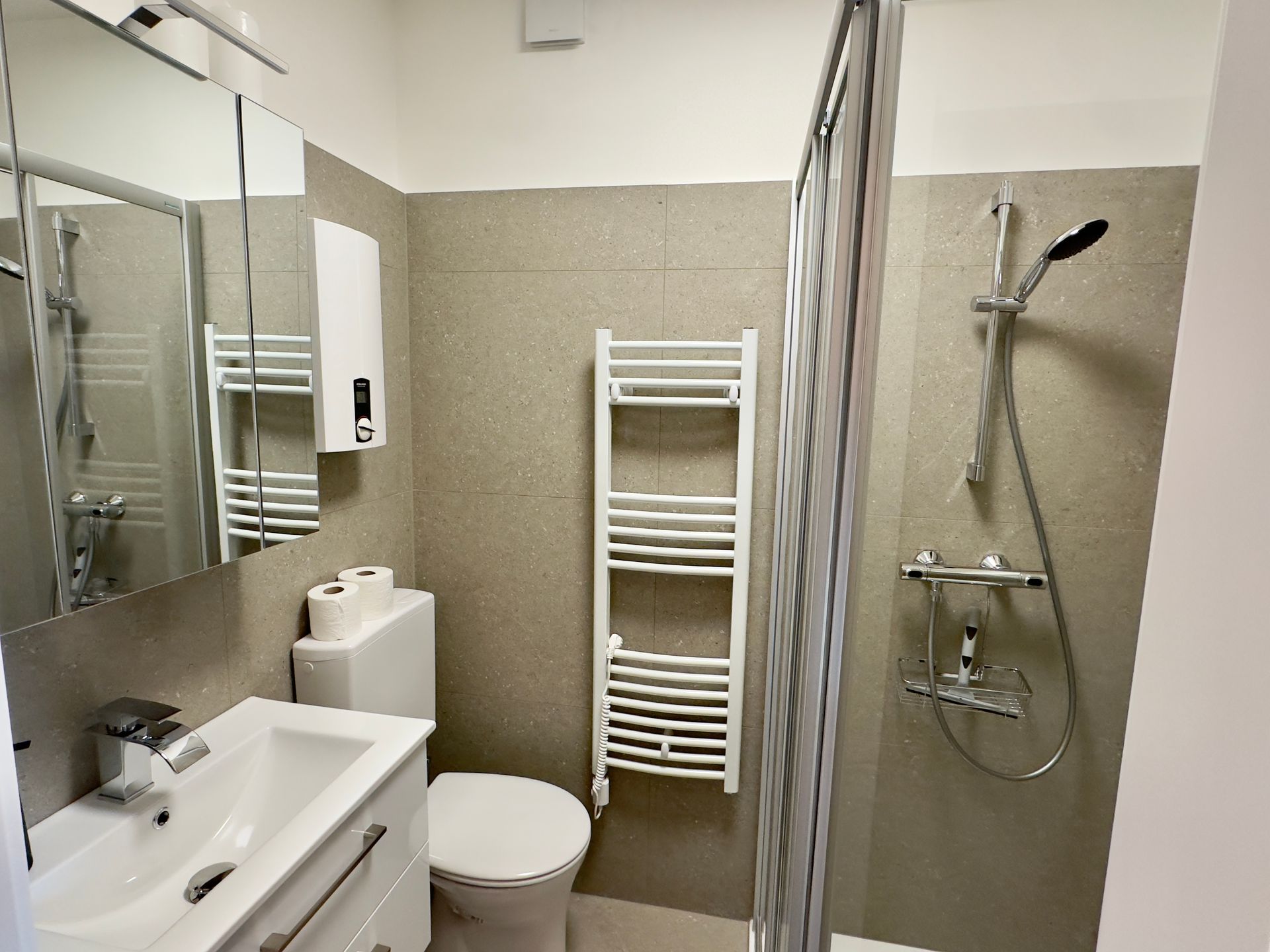 Kleines Badezimmer mit Waschbecken, Toilette, Handtuchhalter, Spiegel und ebenerdiger Dusche.