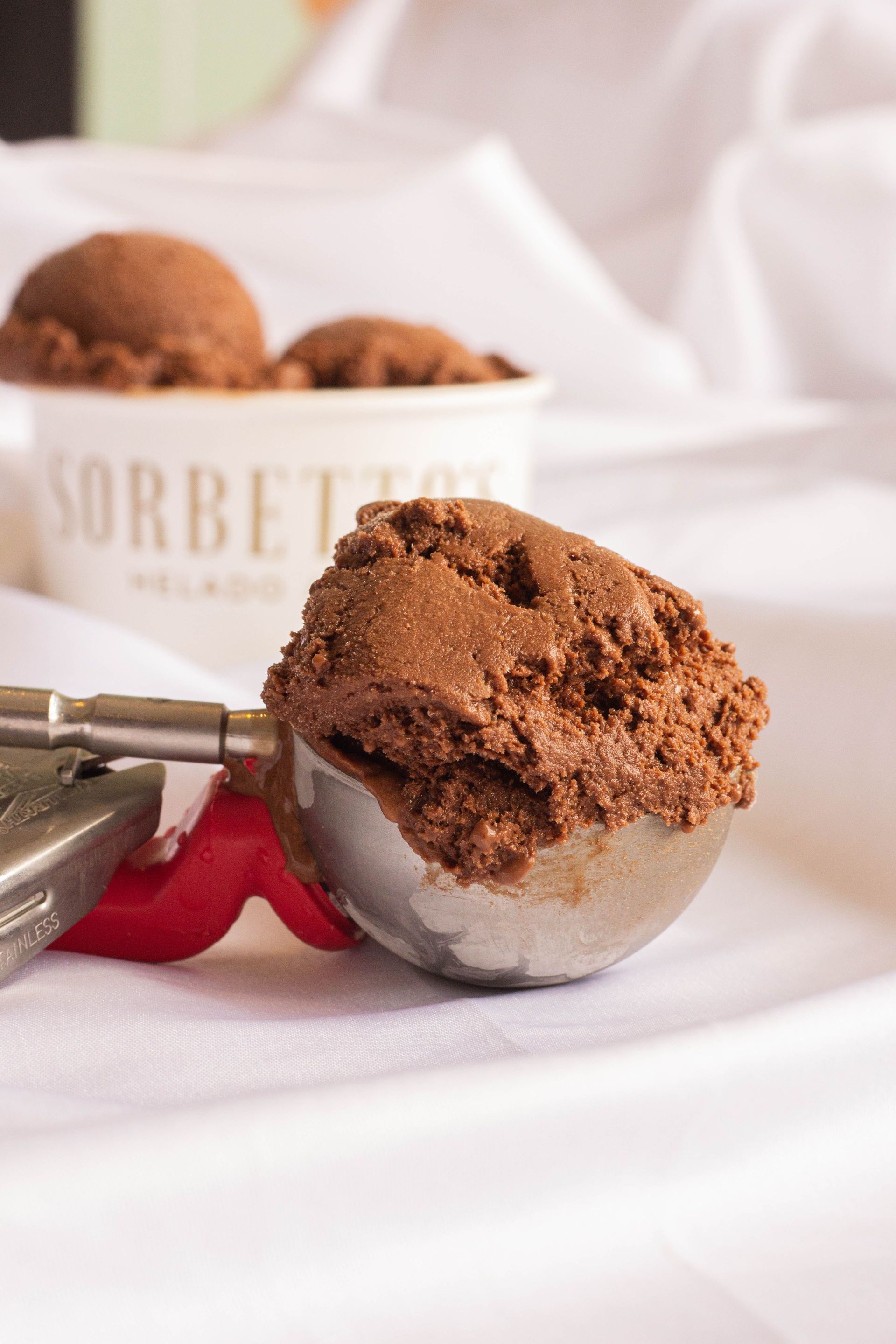 Sorbete de chocolate en una cuchara metálica con mango rojo sobre una mesa blanca, con un recipiente de sorbete al fondo.