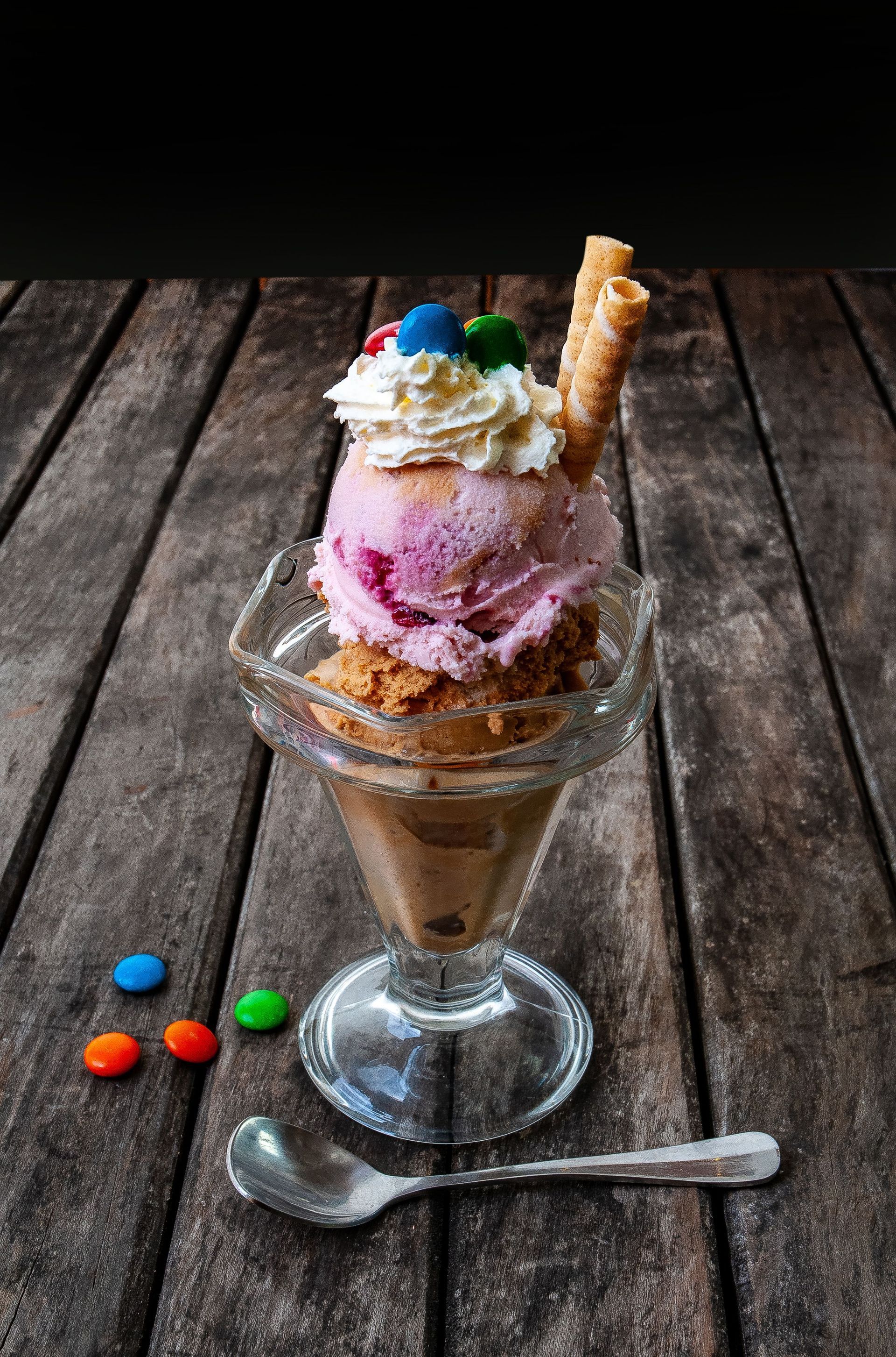 Helado tipo sundae en vaso sobre una mesa rústica de madera, coronado con crema batida y barquillo, con cuchara y caramelos.