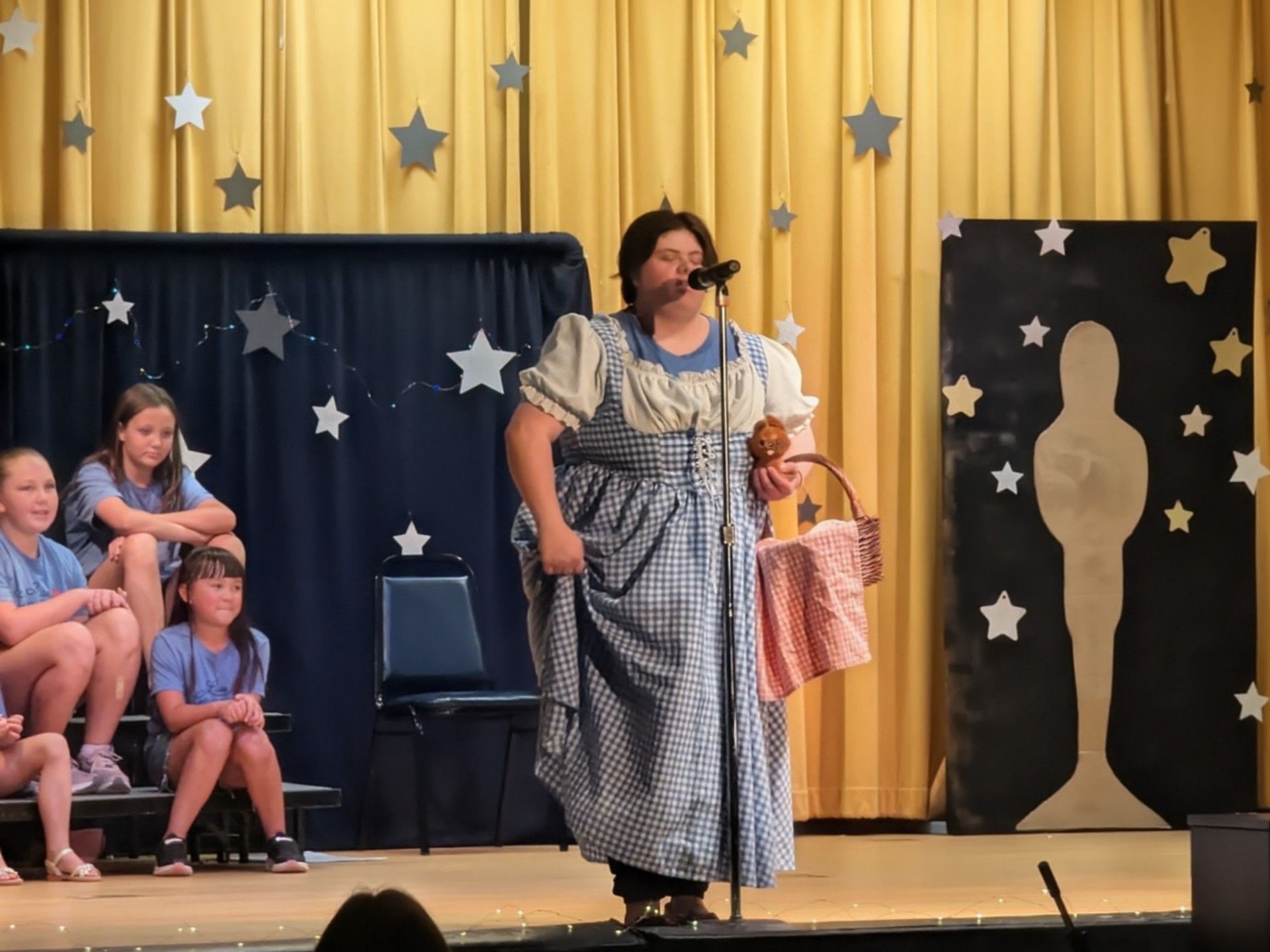 Person in blue plaid dress on stage with microphone, starry backdrop, seated children at left