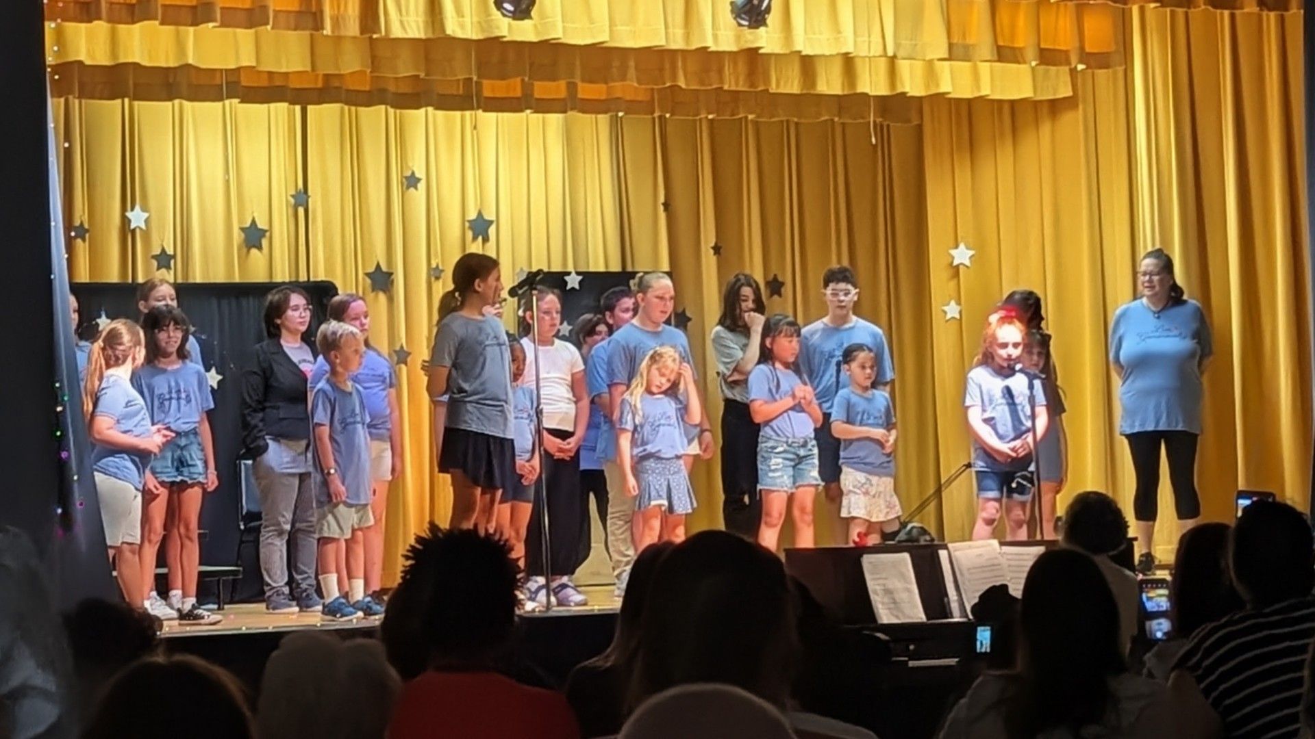 Children singing on a school stage with yellow curtains and microphones, watched by an audience