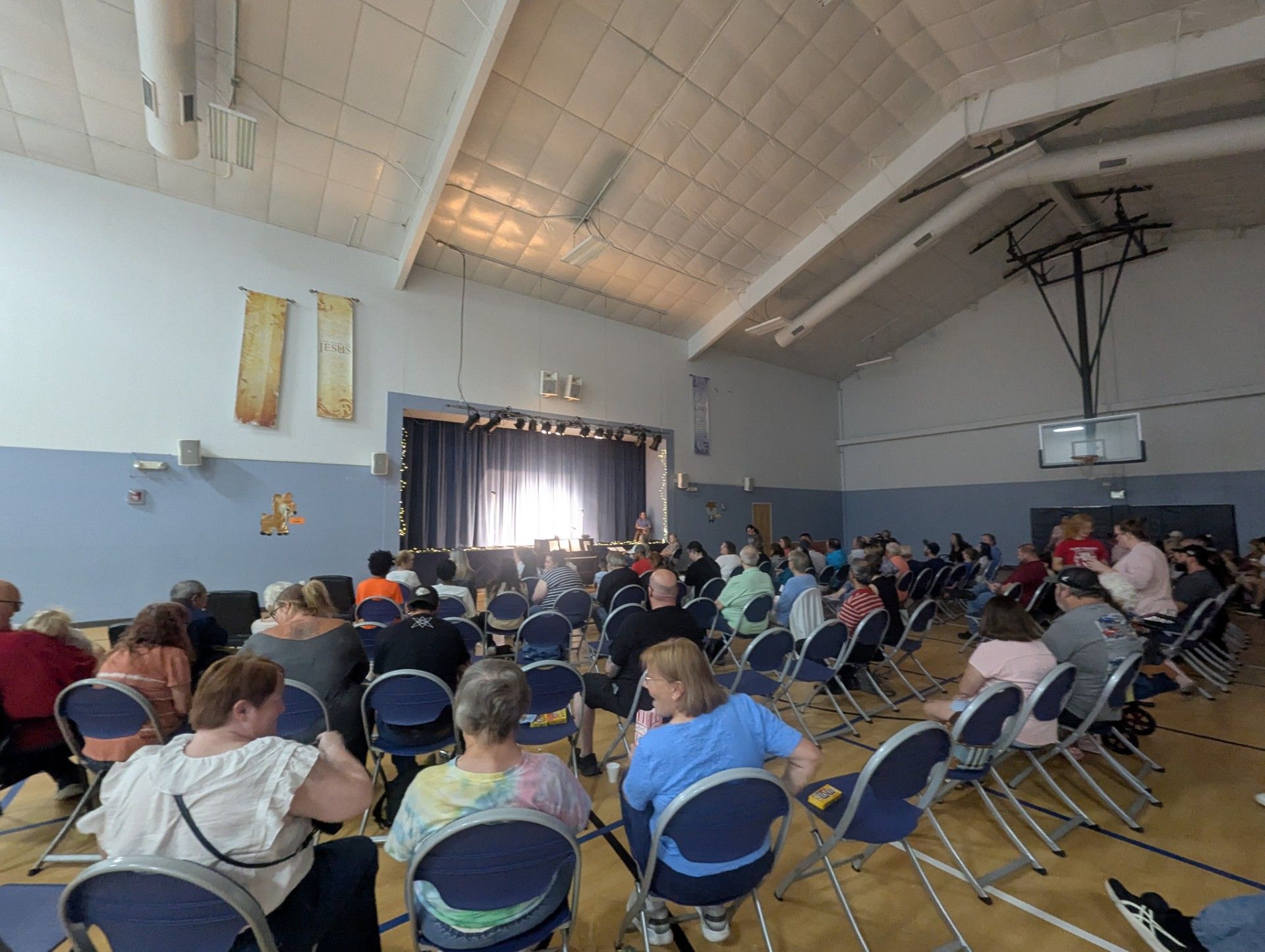 Audience seated in a school gym watching a presentation on a stage with a curtain backdrop