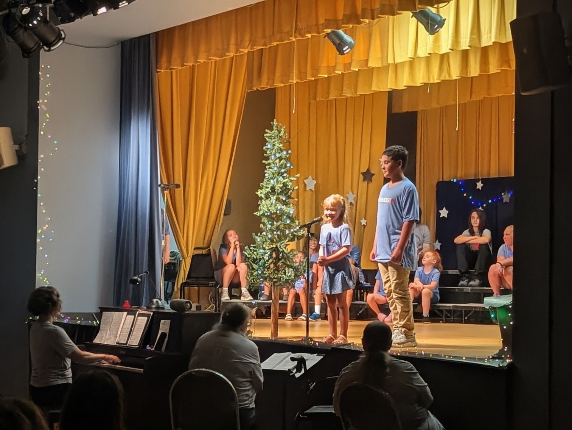 Children performing on a stage with gold curtains, a lit tree, and seated audience members in front.