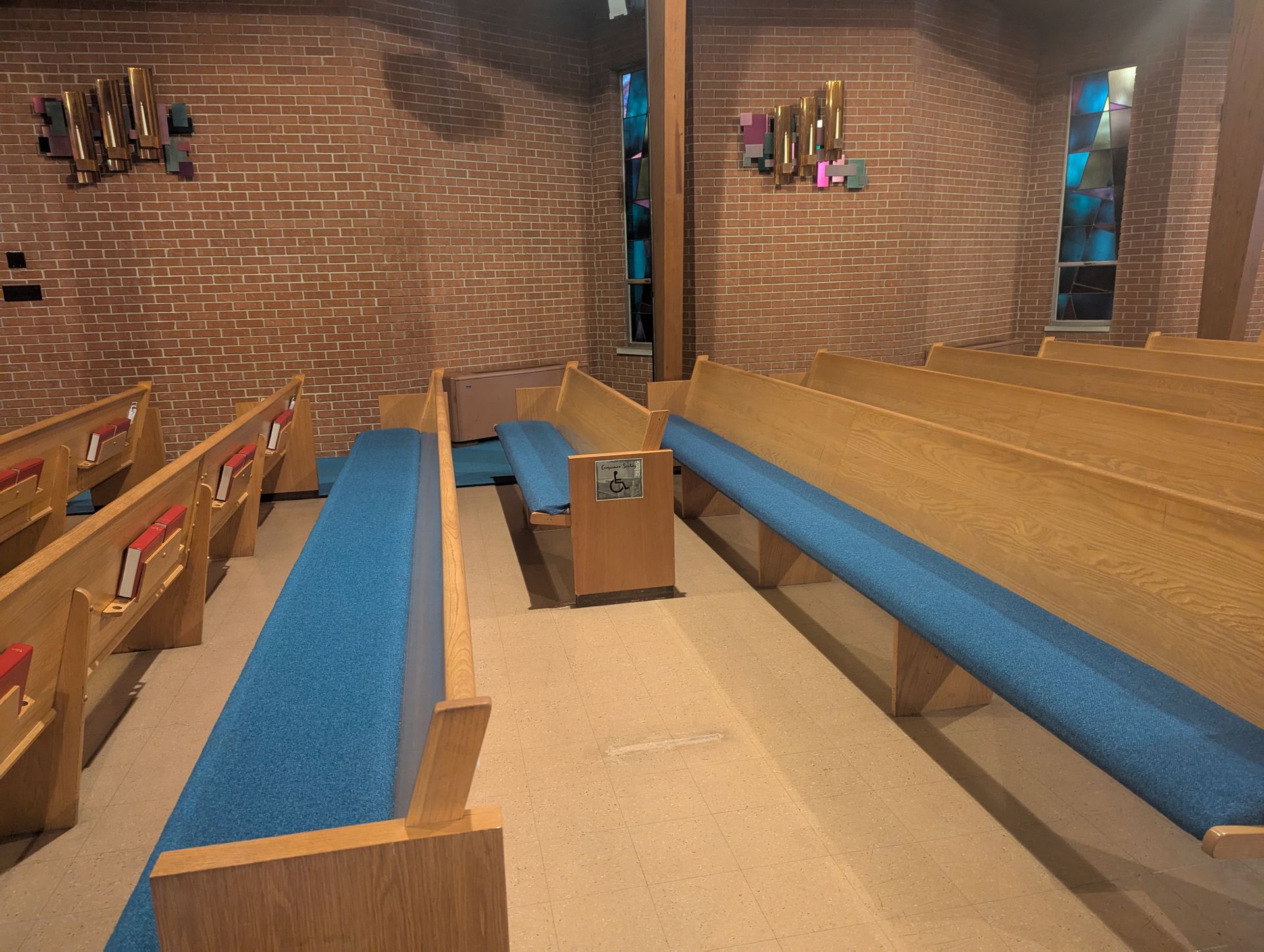 Empty church interior with blue-cushioned wooden pews and a pulpit against brick walls.