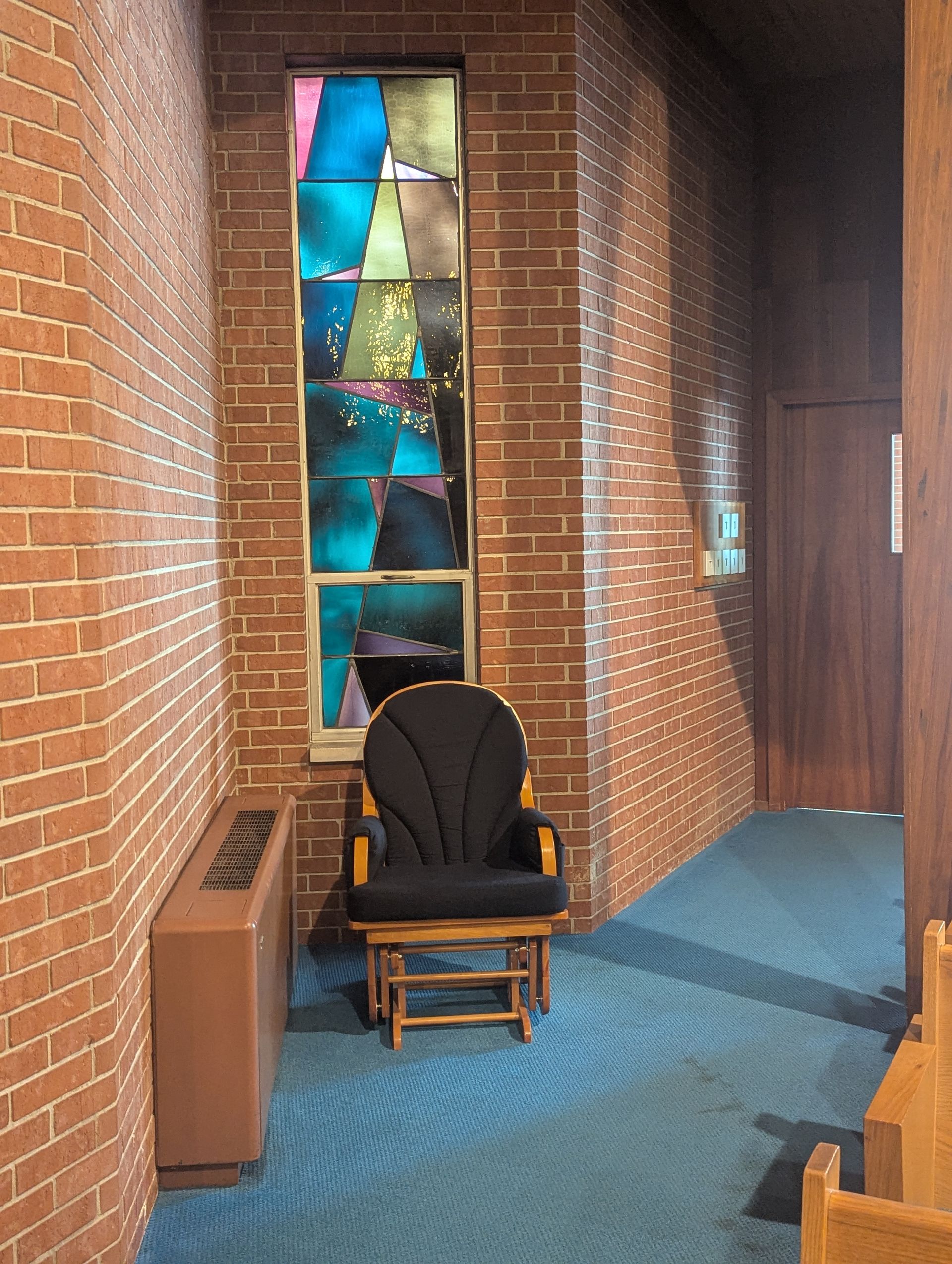 Brick hallway with stained-glass window, black chair, and blue carpet leading to a dark wooden door.