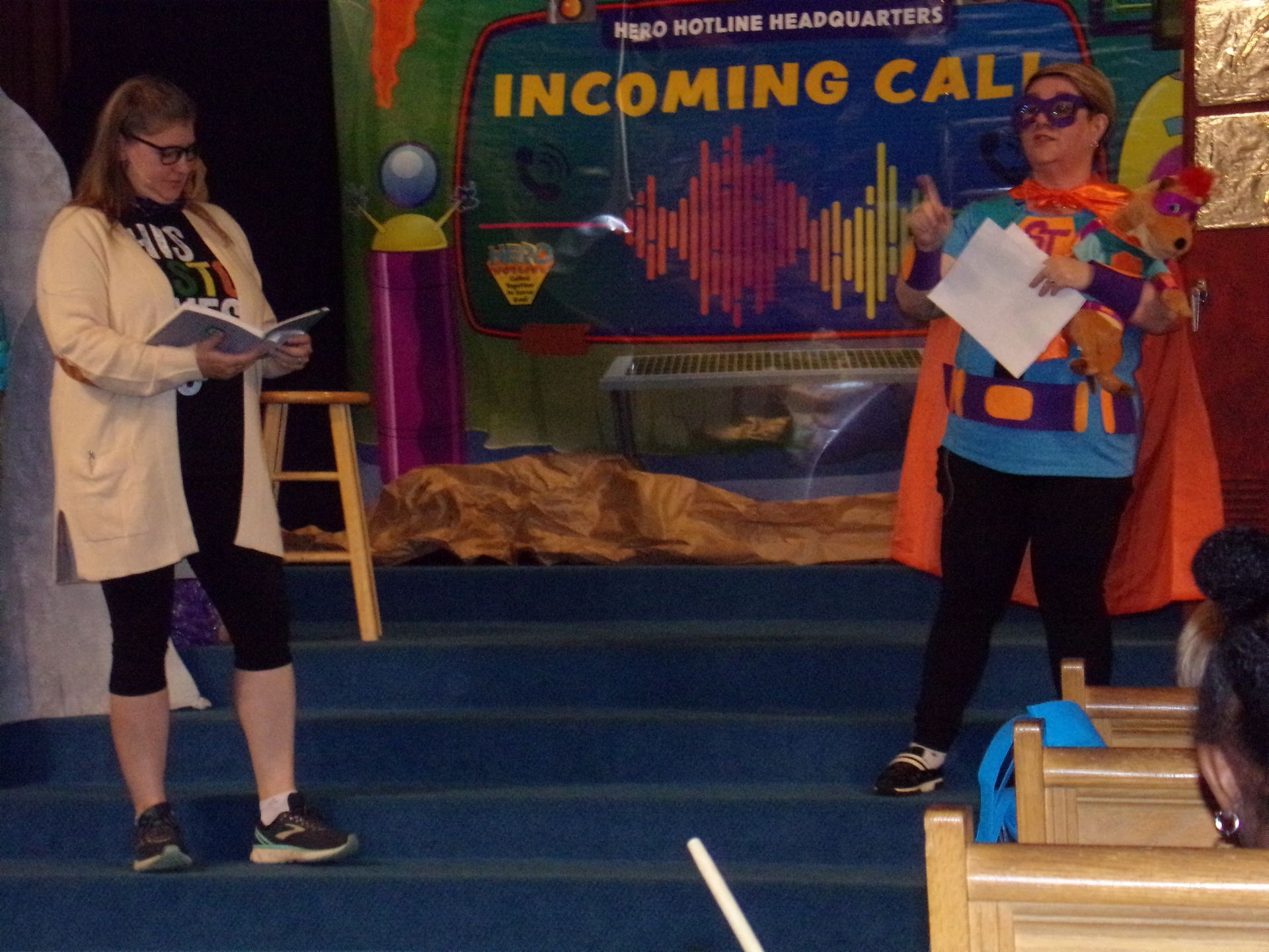Two women on a small stage beneath an “Incoming Call” sign, one holding papers, one at a microphone.