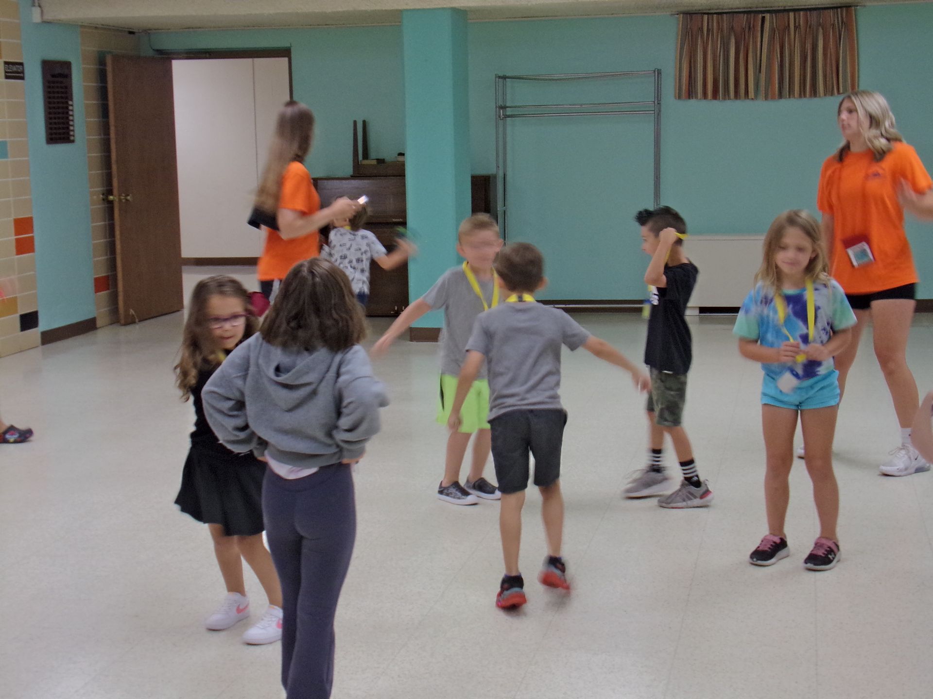 Children dancing in a classroom, with two adults supervising in the background.