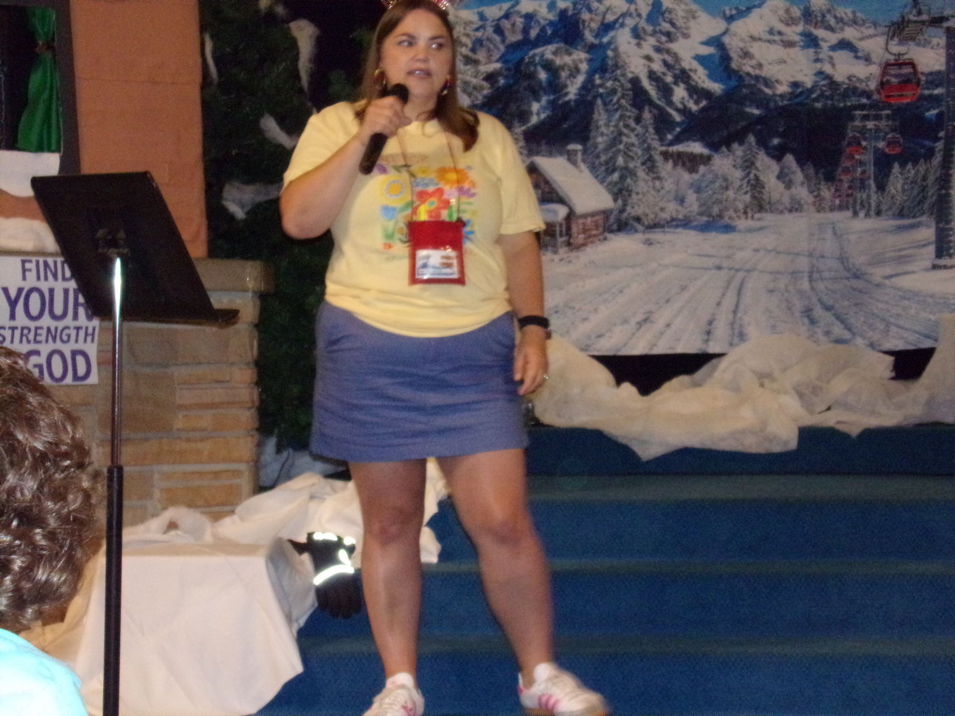 Woman speaking into a microphone on a stage with a snowy mountain backdrop.