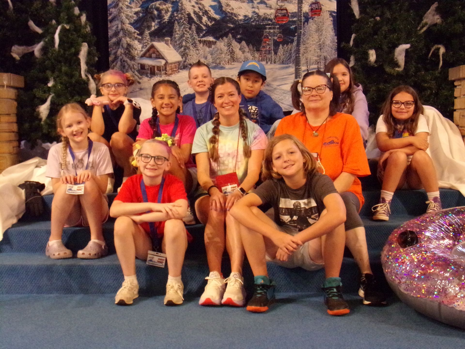 Group of smiling children posing on a blue stage in front of a decorated holiday backdrop.