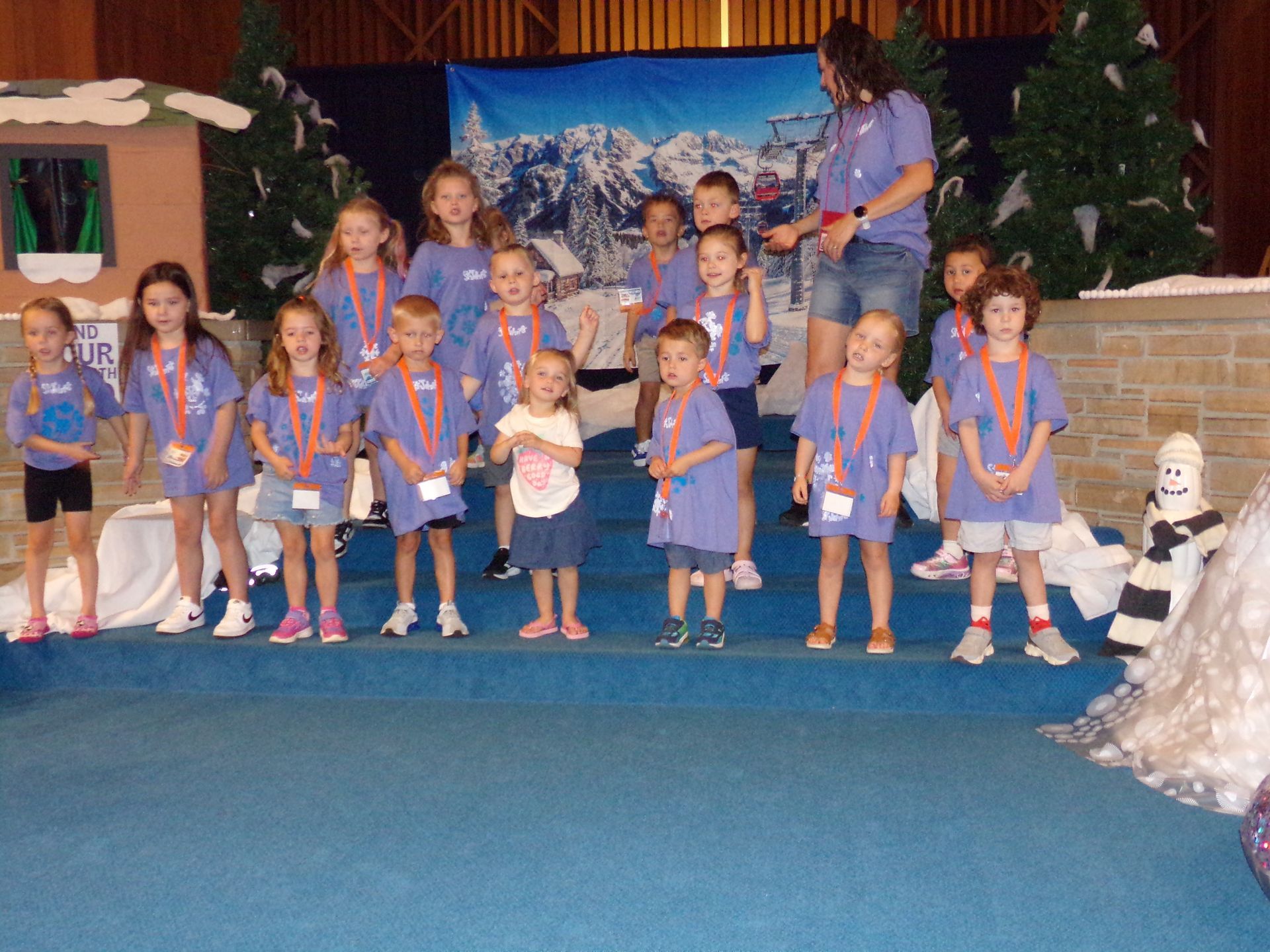 Children in blue outfits stand on a stage at a school performance with Christmas decorations.